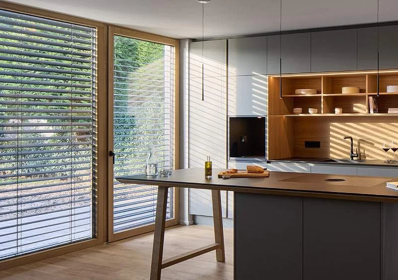 Moderne Küche mit großen Fenstern, Holzregalen und einem Küchenblock, die Sonne fällt durch die Jalousien.