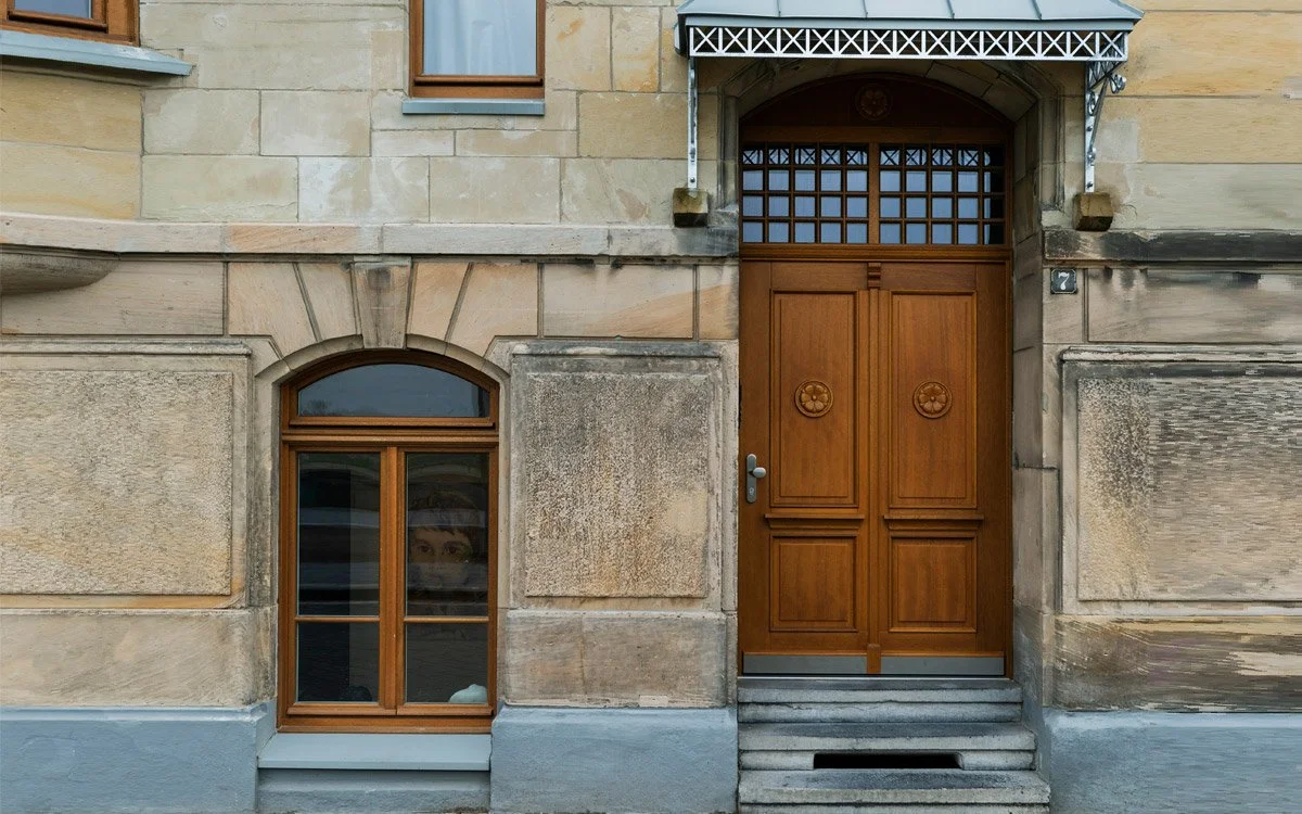 Ein Gebäudefront mit einer Holztür und einem Fenster darunter, alles in einem steinernen Rahmen, im oberen Bereich befindet sich ein weiteres kleines Fenster.