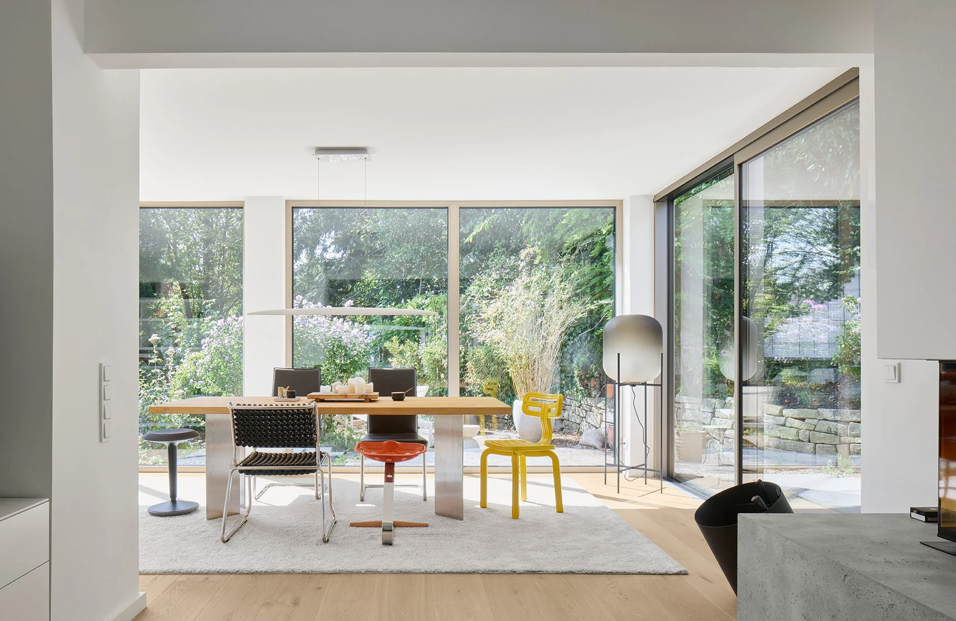 Moderner Essbereich mit großem Tisch, Stühlen und Blick nach draußen auf einen Garten mit Bäumen und Blumen, große Glasfenster und helle Innenraumgestaltung.