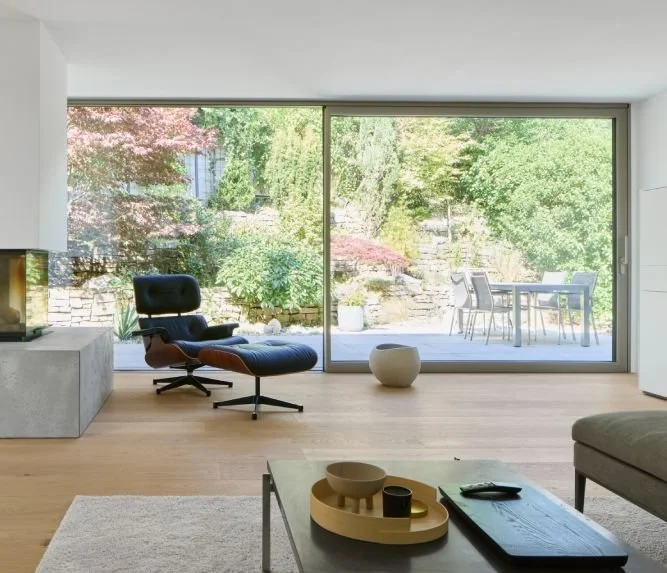 Modernes Wohnzimmer mit großen Aluminium Schiebe-Hebe-Fenstern, Blick auf einen Garten mit Terrasse, Gartenmöbel und viel Grün.