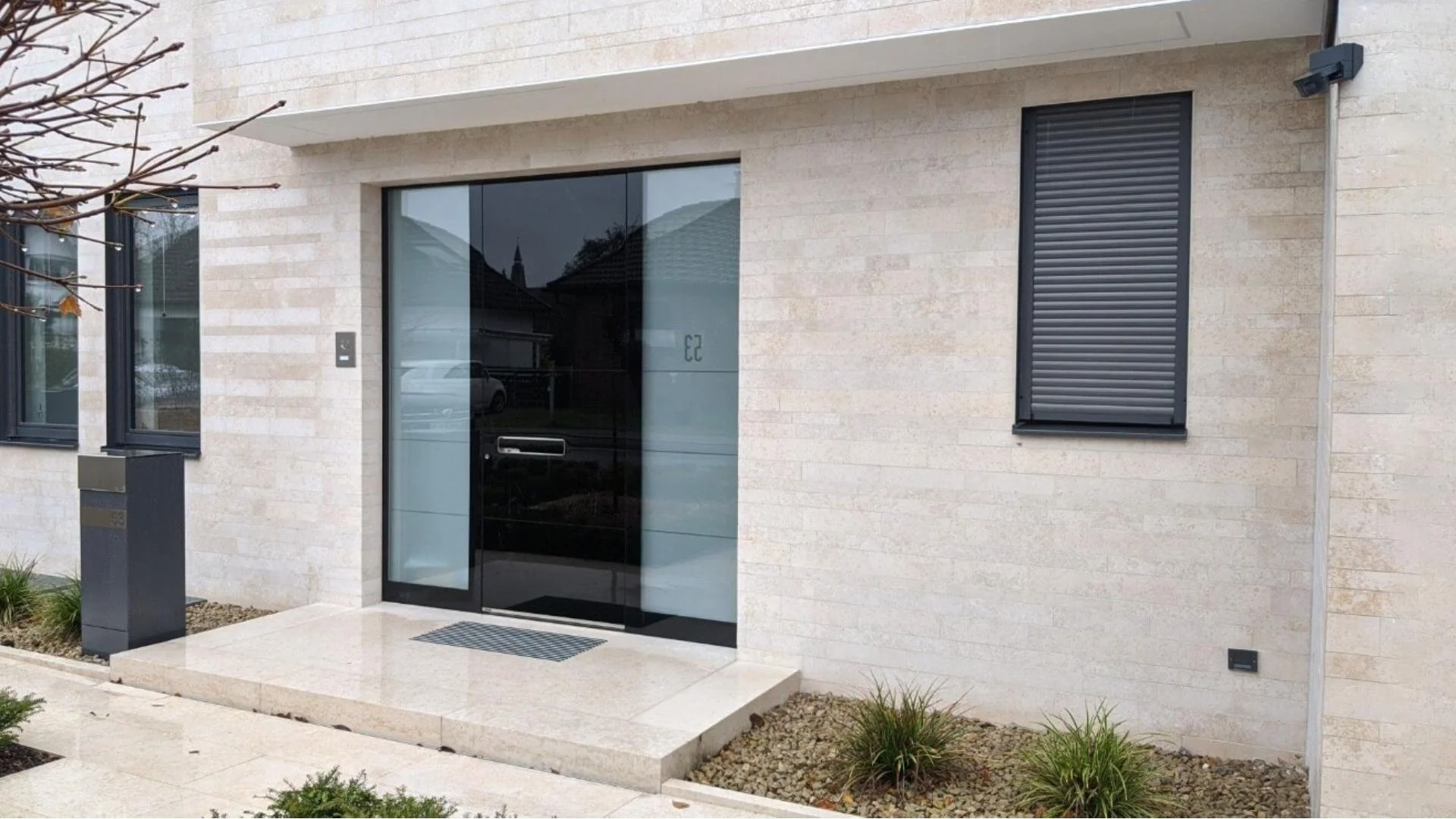 Moderne Hausfassade mit Haustür aus Glas, schwarzen Rollläden an den Fenstern und kleinen Garten mit Pflanzen.