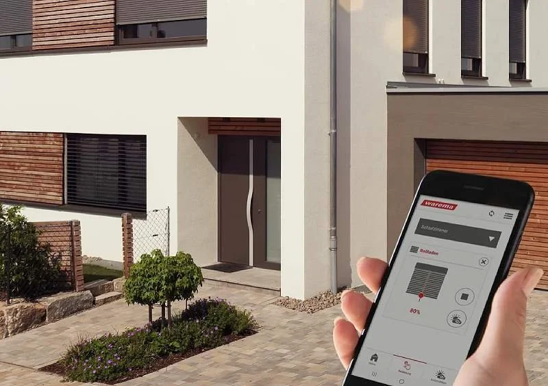 Moderne Fassade mit Raffstoren mit smartem Steuerungssystem auf Smartphone im Vordergrund, modern gebautes Haus mit Garten und Zufahrt.
