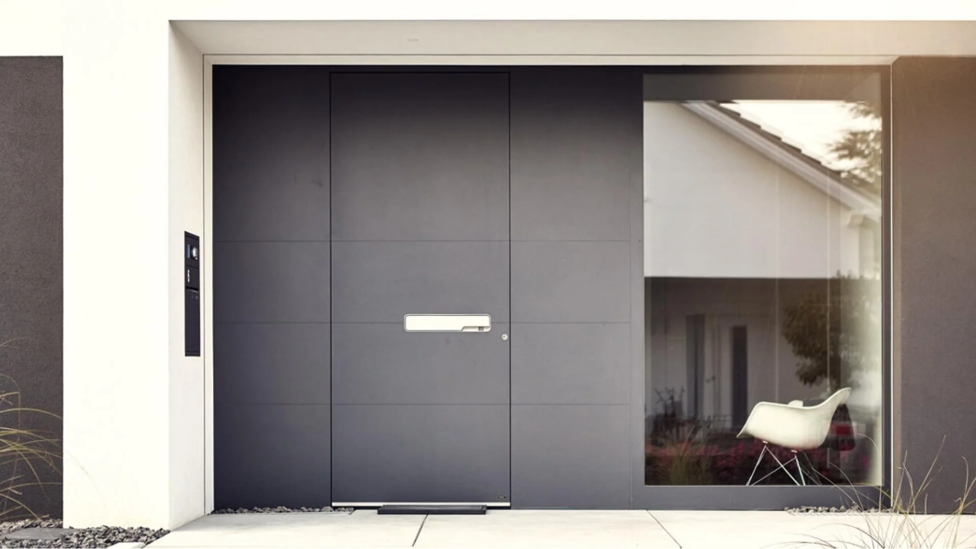 Moderne Eingangstür mit schwarzer Oberfläche, seitlicher Glasfenster und einem weißen Stuhl im Eingangsbereich. Reflektion eines Hauses im Glas.