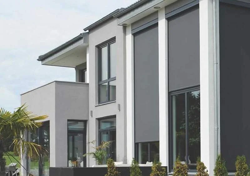 Modernes zweistöckiges Haus mit großen Fenstern und Senkrechtmarkisen, umgeben von Garten mit Palmen und Büschen.