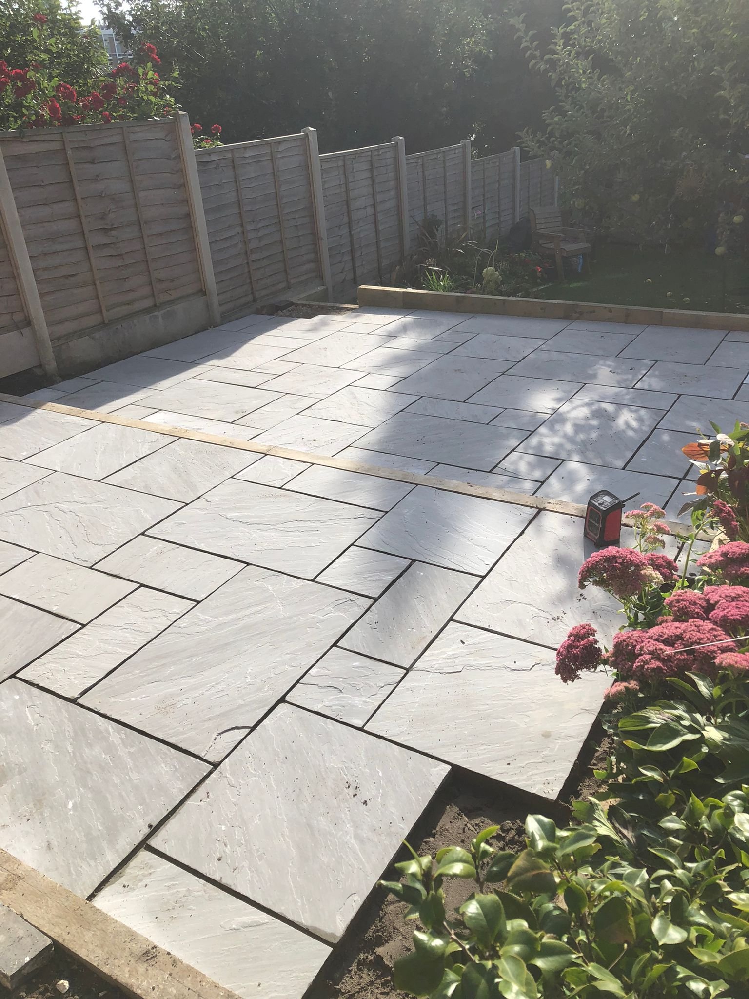 A backyard patio with newly installed large white stone tiles, surrounded by a wooden fence, garden plants, and pink flowers.