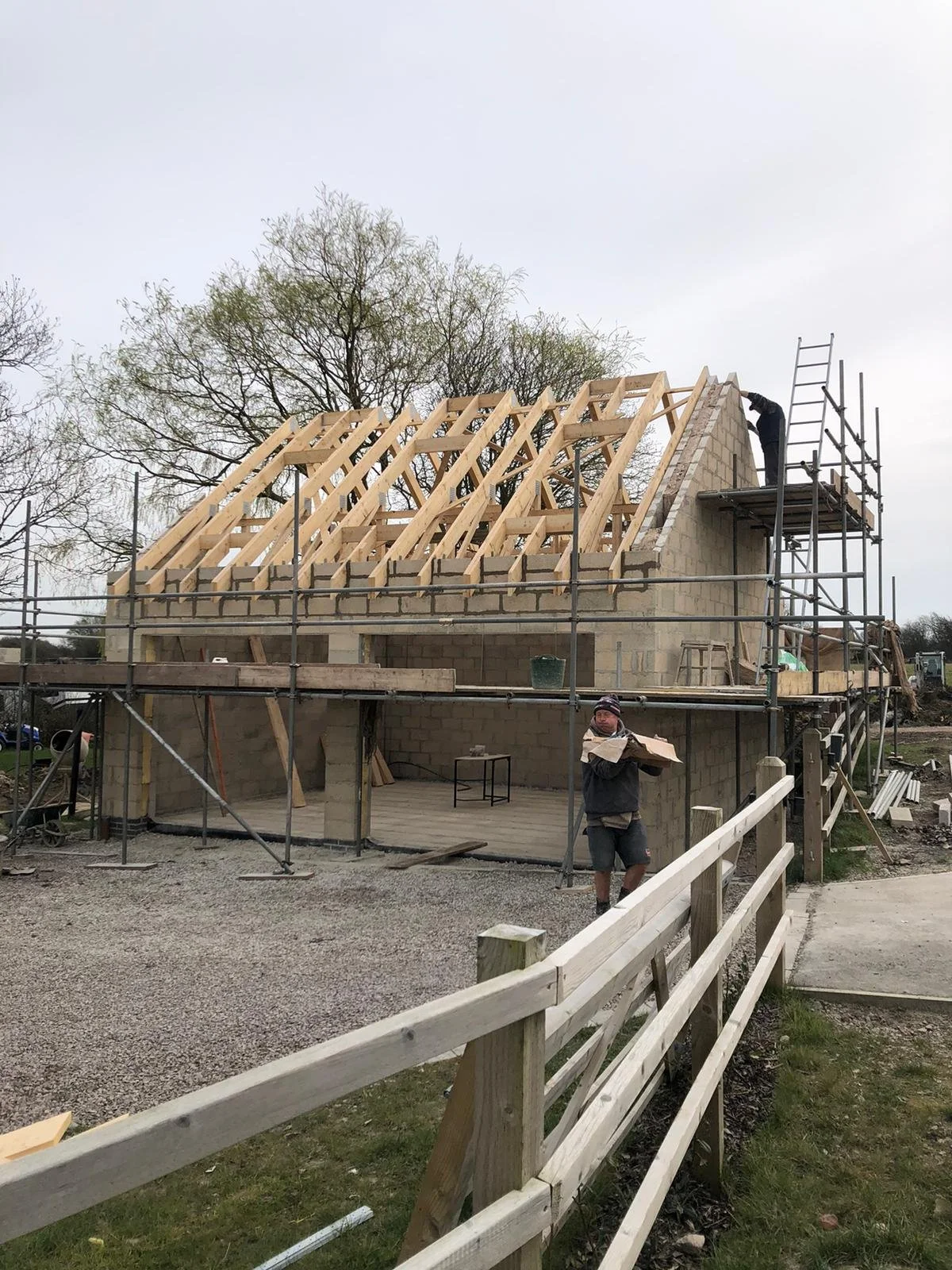 Construction workers building a house with a wooden roof frame and scaffolding, with trees in the background.