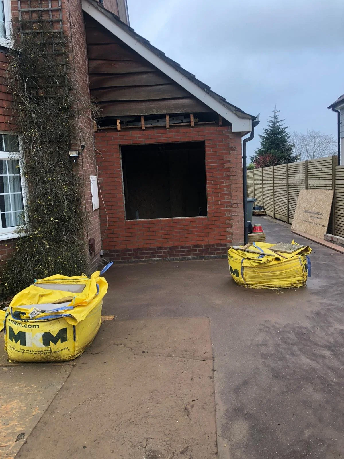 Construction site of a house with a brick exterior, window, and an unfinished opening in the wall. Two large yellow bags of construction materials are on the ground, and there is a wooden fence on the right side of the scene.