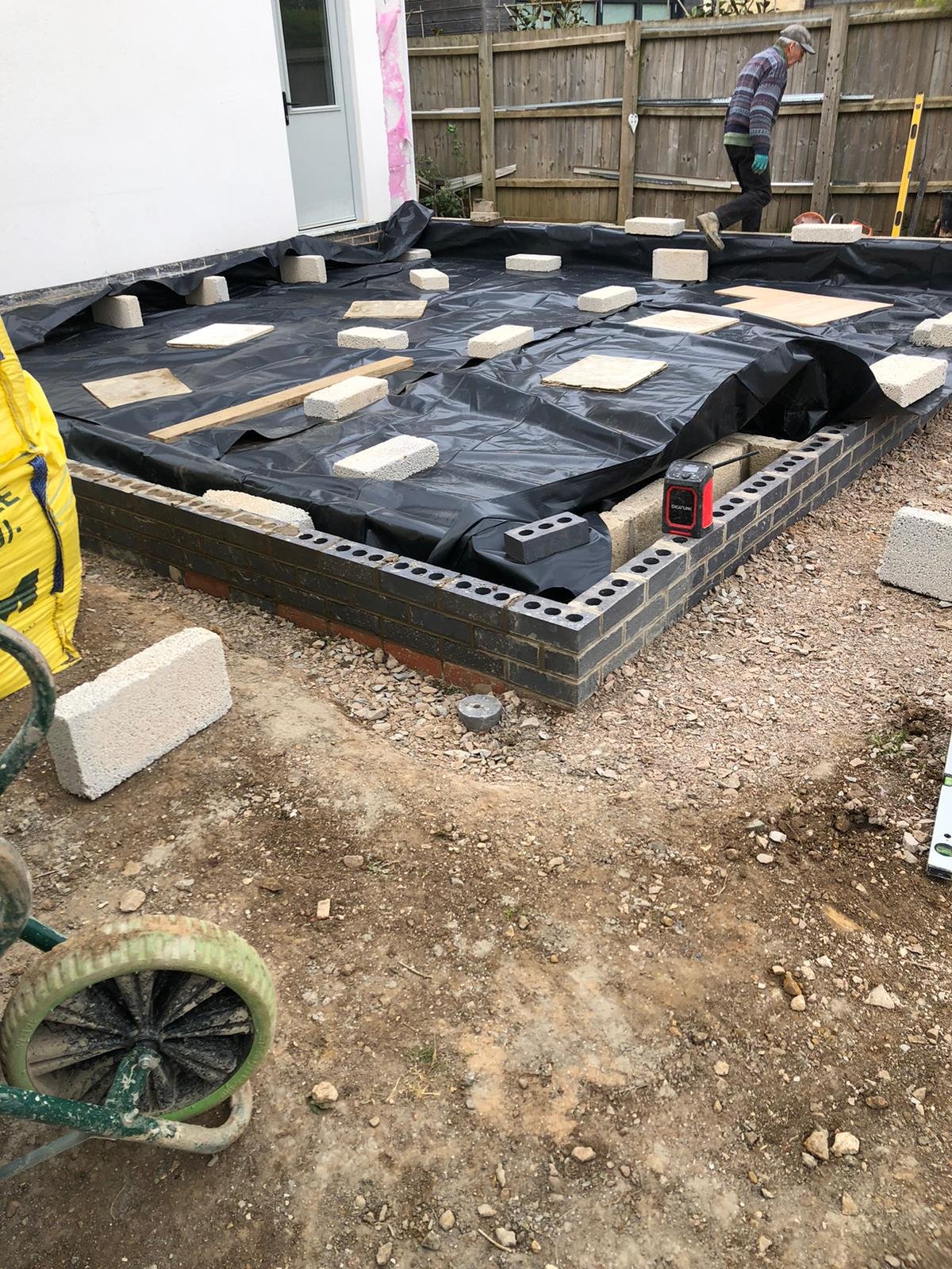 Grantham builder G East Builder. working on a construction site laying bricks for a building foundation, with black plastic sheeting and cinder blocks in place.