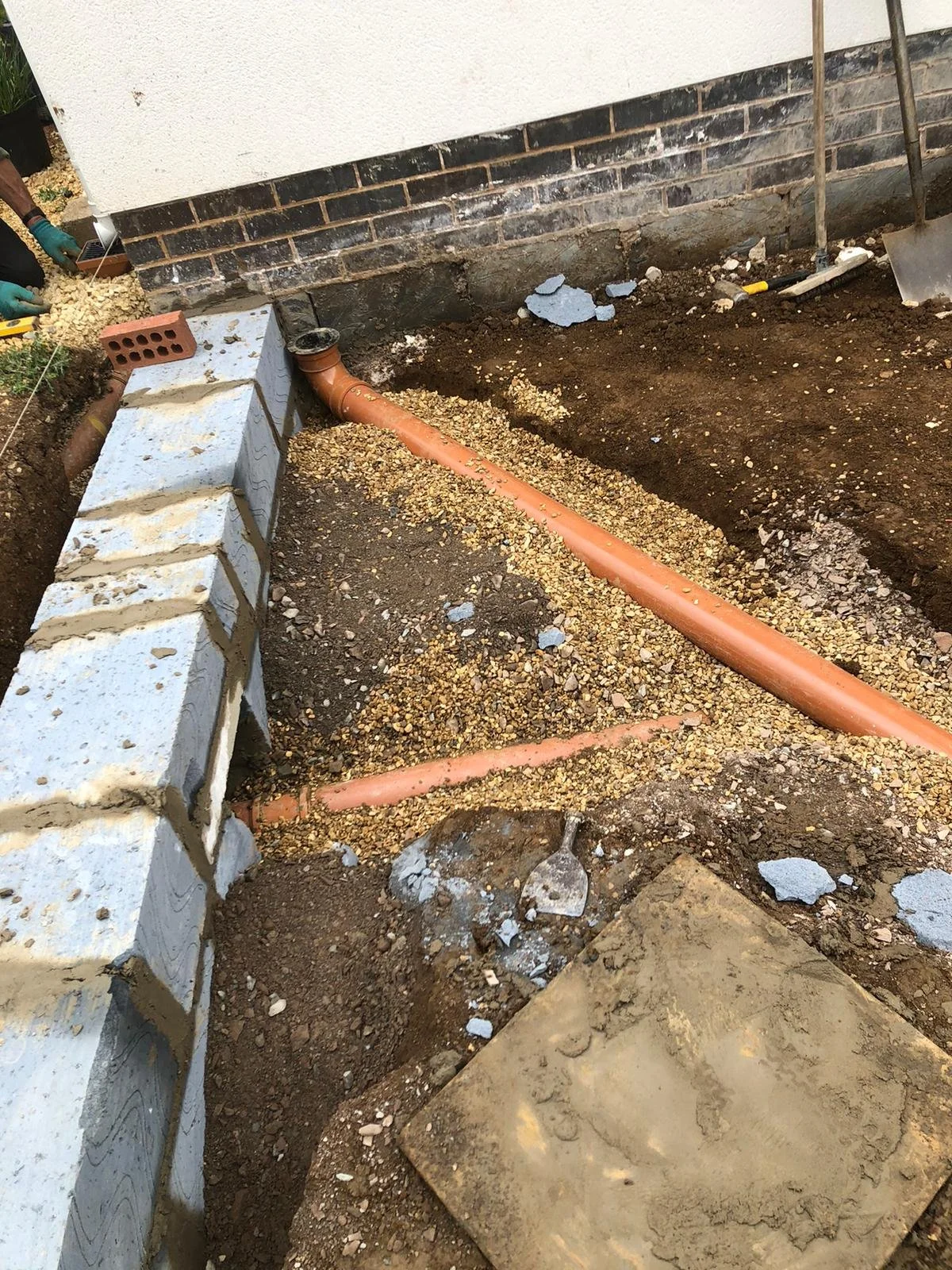 Construction site with exposed orange plumbing pipe, gravel, and bricks near a house foundation.