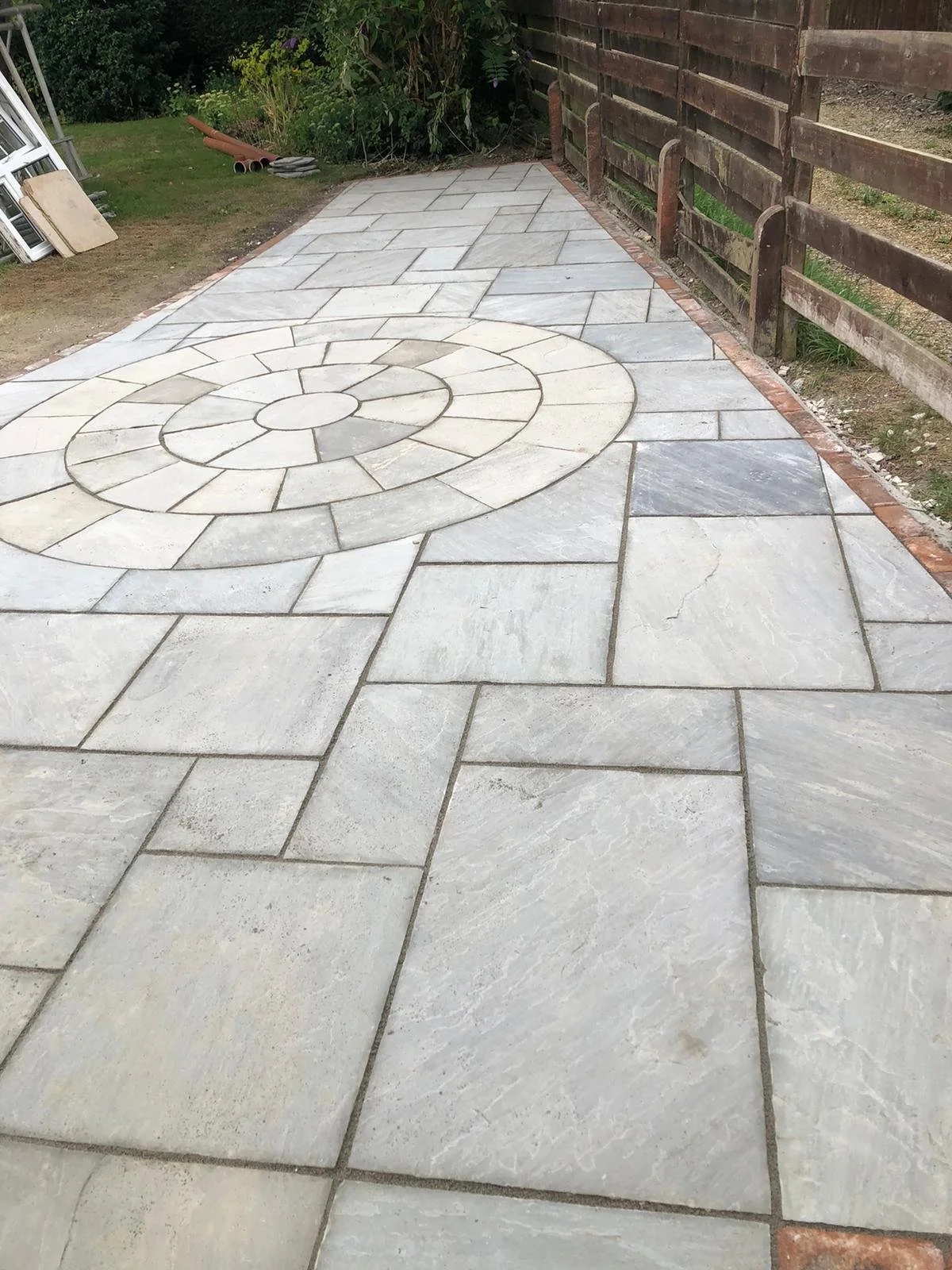 Newly paved stone patio with a circular pattern feature in the center, adjacent to a wooden fence and garden area.