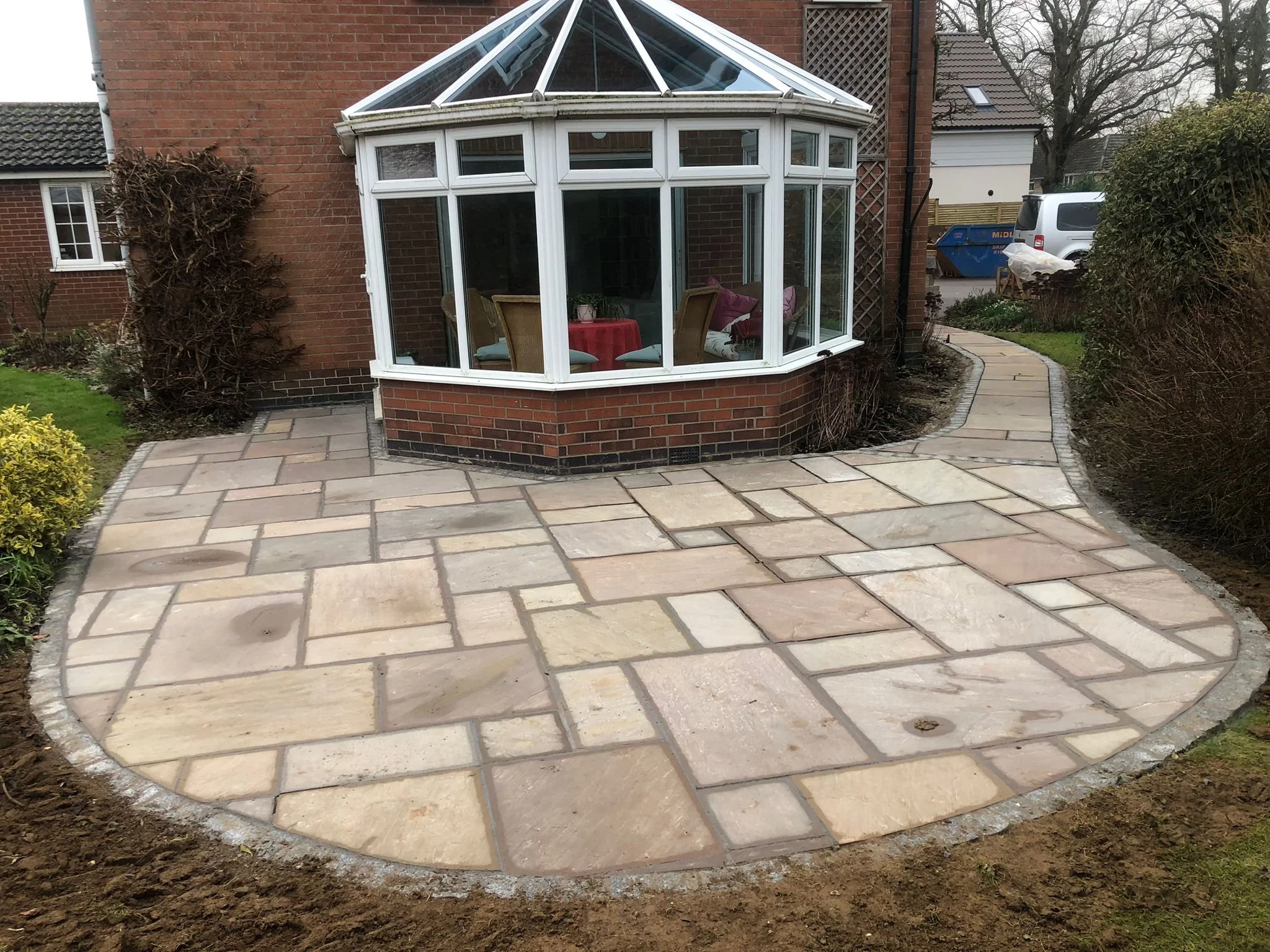 Newly paved stone patio with a curved shape surrounds a brick house with a glass roofed conservatory. The patio has several large, rectangular light-colored stones.