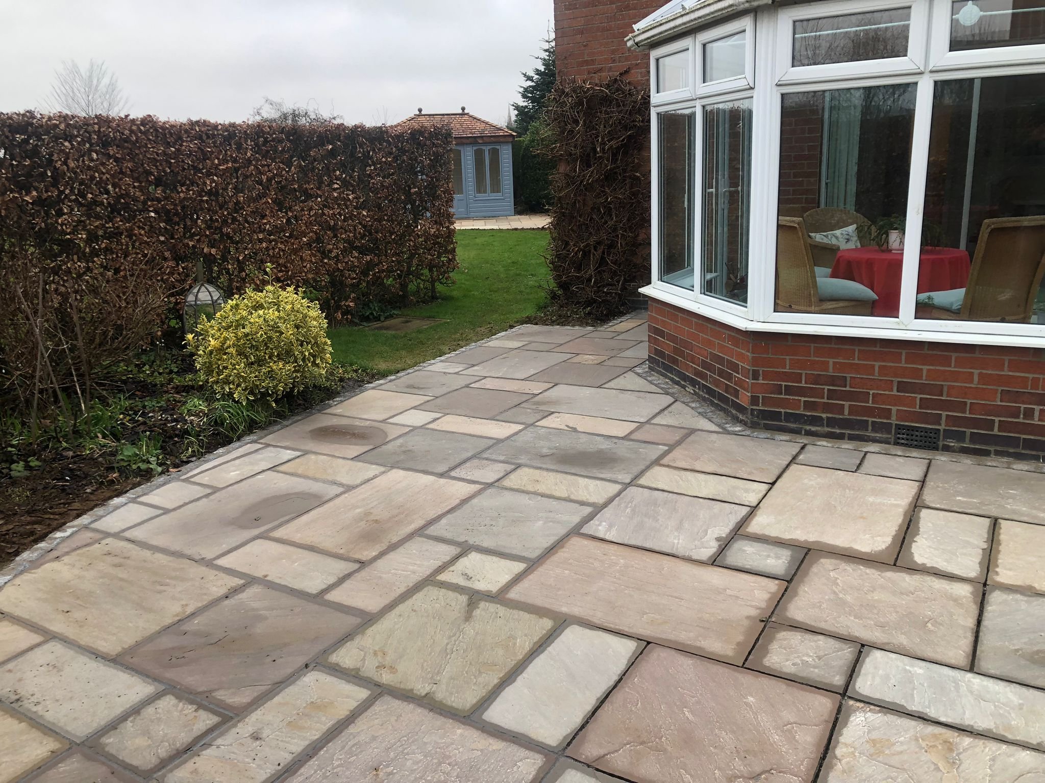 A paved backyard patio area with cream-colored stone tiles, a small garden bed with plants and a decorative lantern, and a brick house with a glass-enclosed room set up for dining.