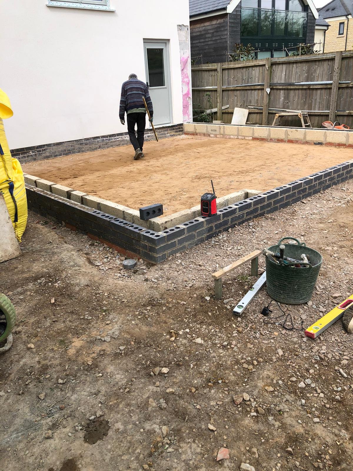 A construction worker walks on a freshly leveled dirt foundation next to a partially built brick wall in a backyard. Construction tools and materials are scattered around, with a bucket of tools in the foreground and neighboring houses in the backgro