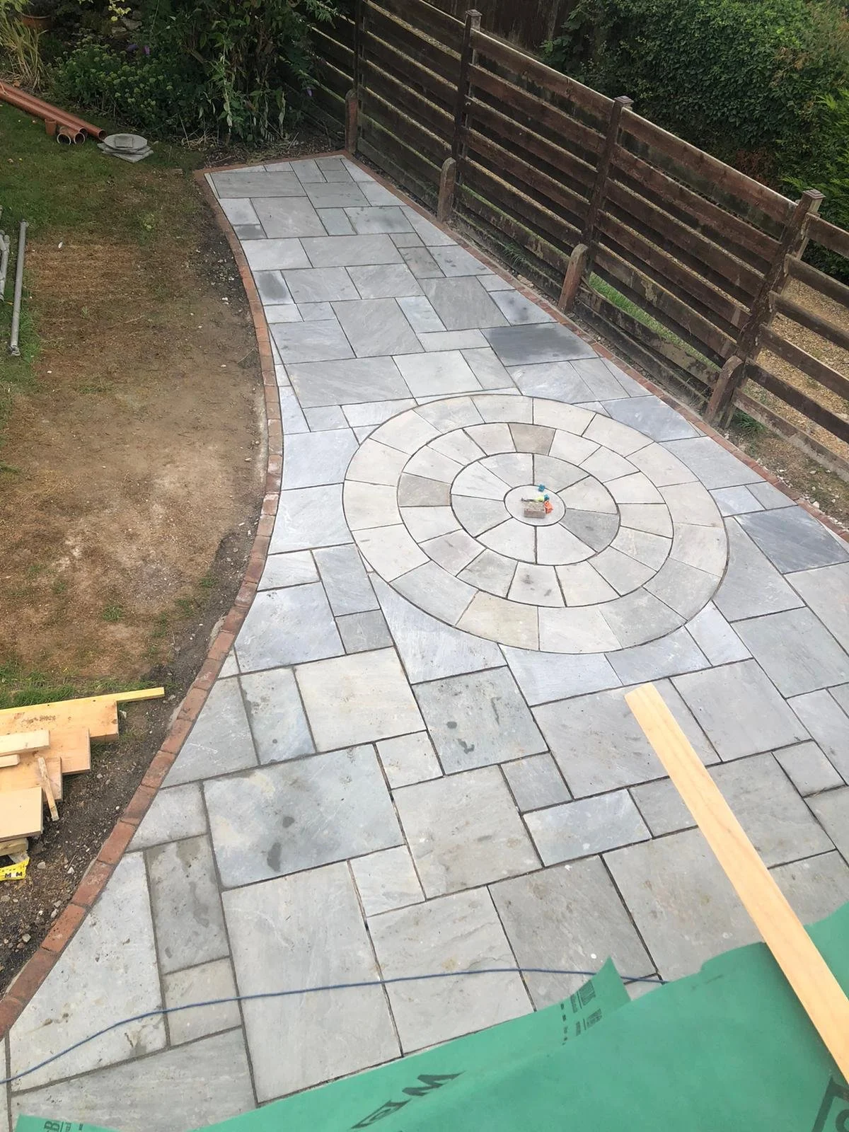Newly paved stone pathway with a circular pattern in the center, bordered by a brick edge, situated next to a grassy area with a wooden fence in the background.
