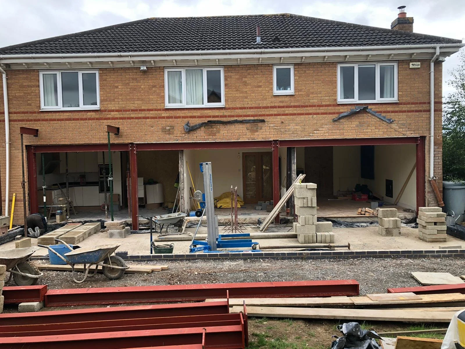 Undergoing home extension with construction materials, bricks, wheelbarrow, and scaffolding in front of a brick house with a tiled roof.
