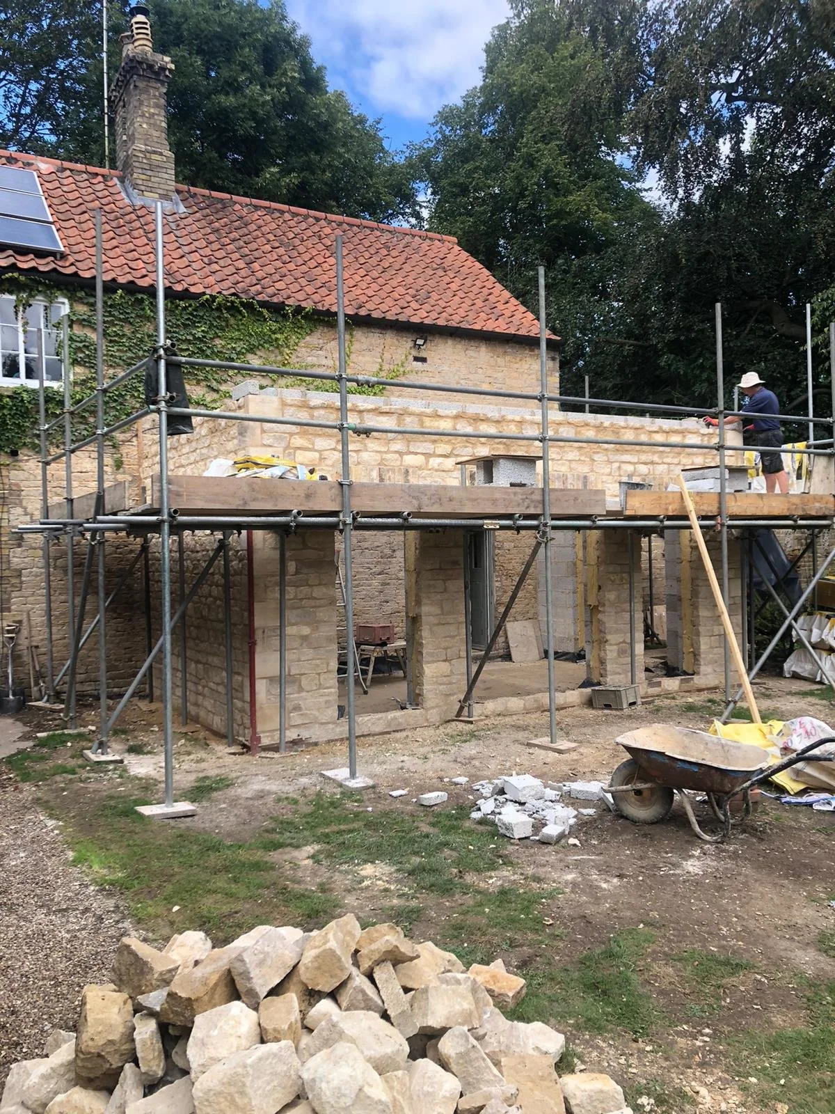 Construction site with scaffolding, brick walls, and a worker wearing a white hat, working on a building extension adjacent to an existing house with a red tiled roof and solar panels.
