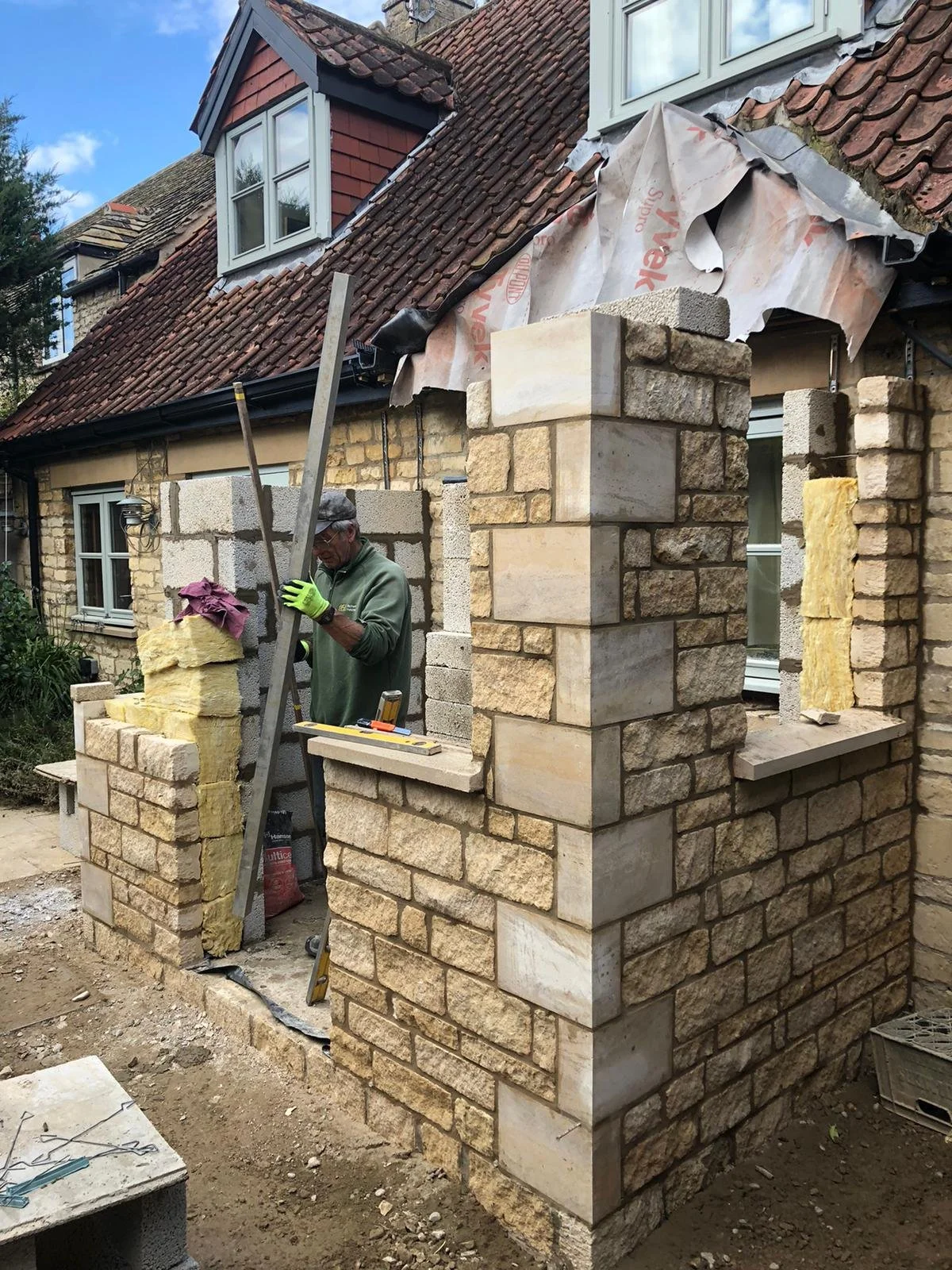A construction worker building a stone wall on a house with a sloped roof and dormer windows, with some insulation and roofing work in progress.