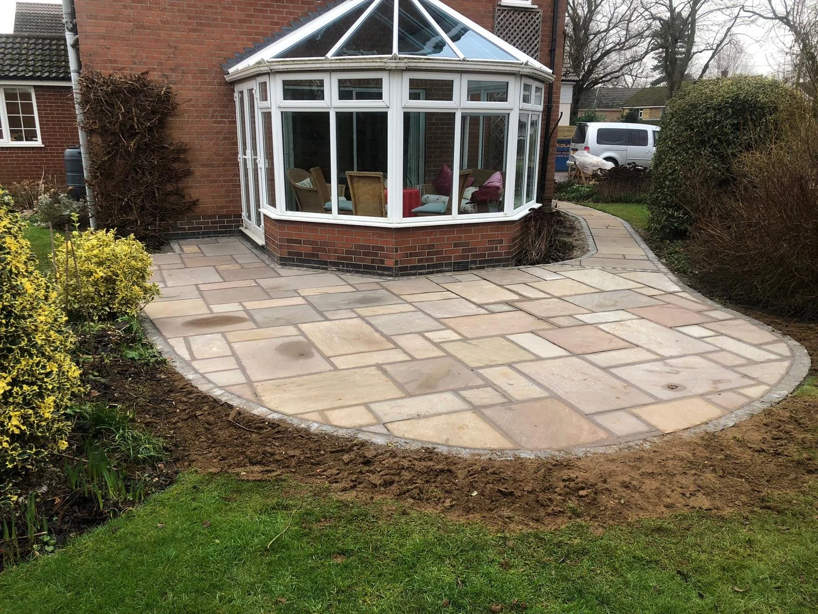 A newly paved stone patio with a curved edge surrounds a brick house with a large glass conservatory. There are bushes and a lawn nearby.