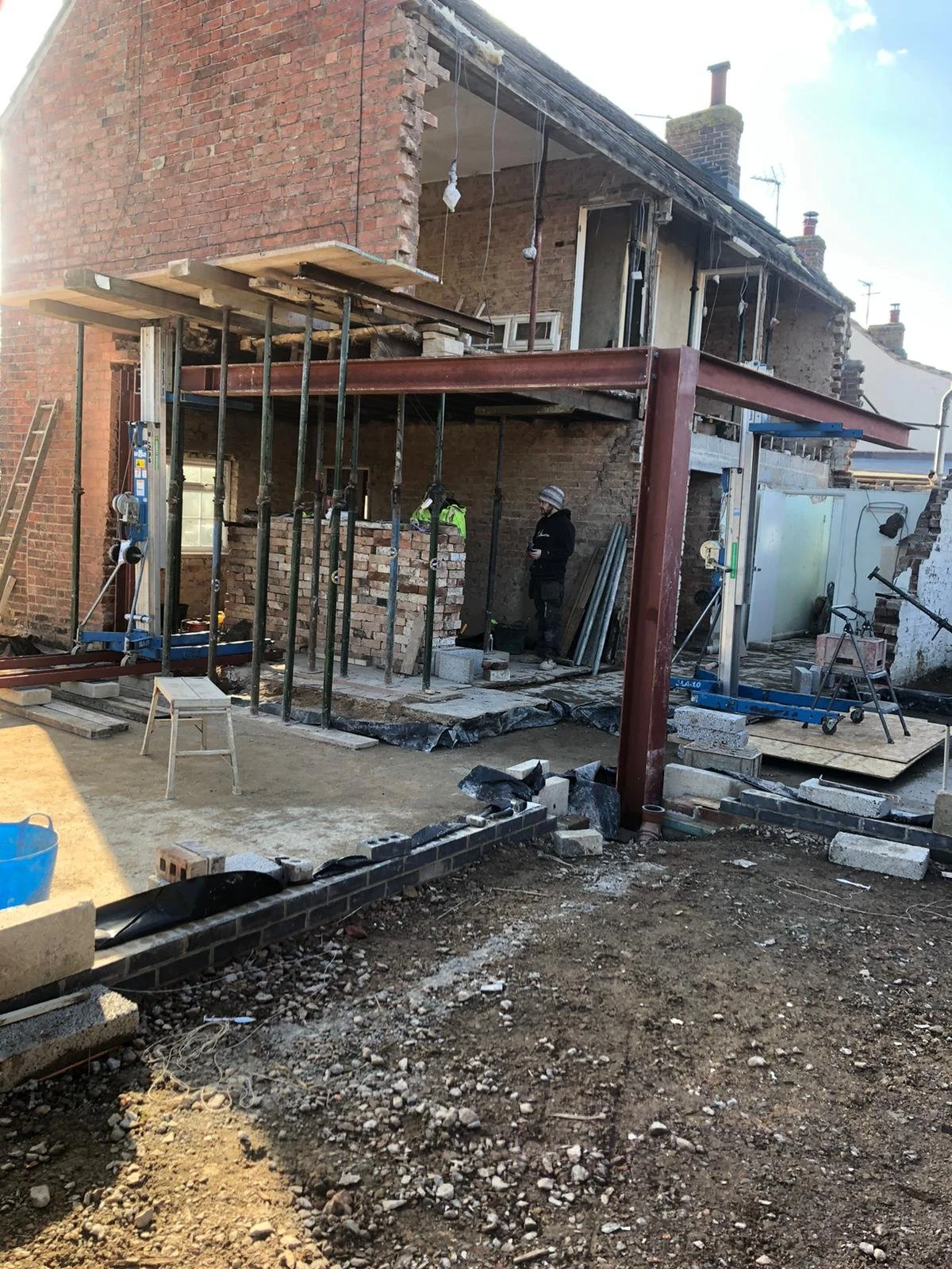 Construction site of a building extension: workers installing brick walls and steel beams on a brick house, with scaffolding and construction tools in the foreground.