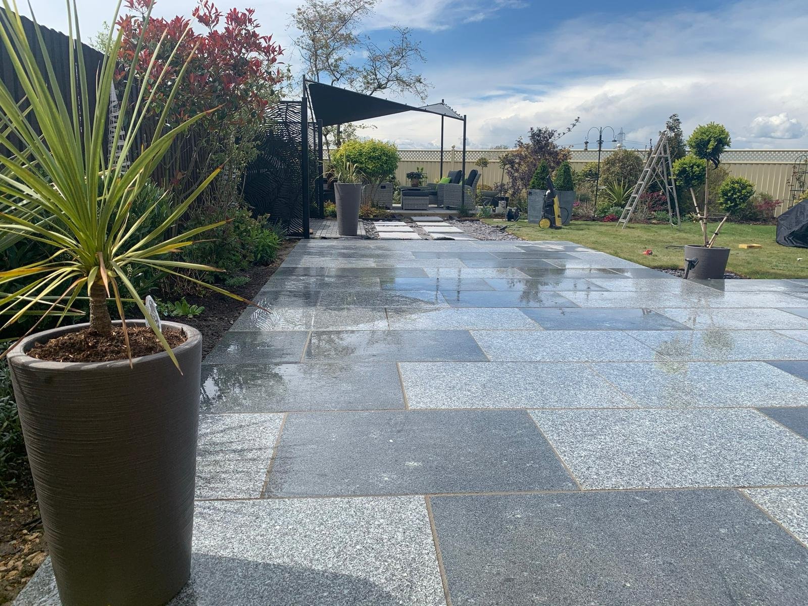 A backyard patio with wet gray stone tiles, potted plants, outdoor furniture, a ladder, and a metal fence, under a partly cloudy sky.