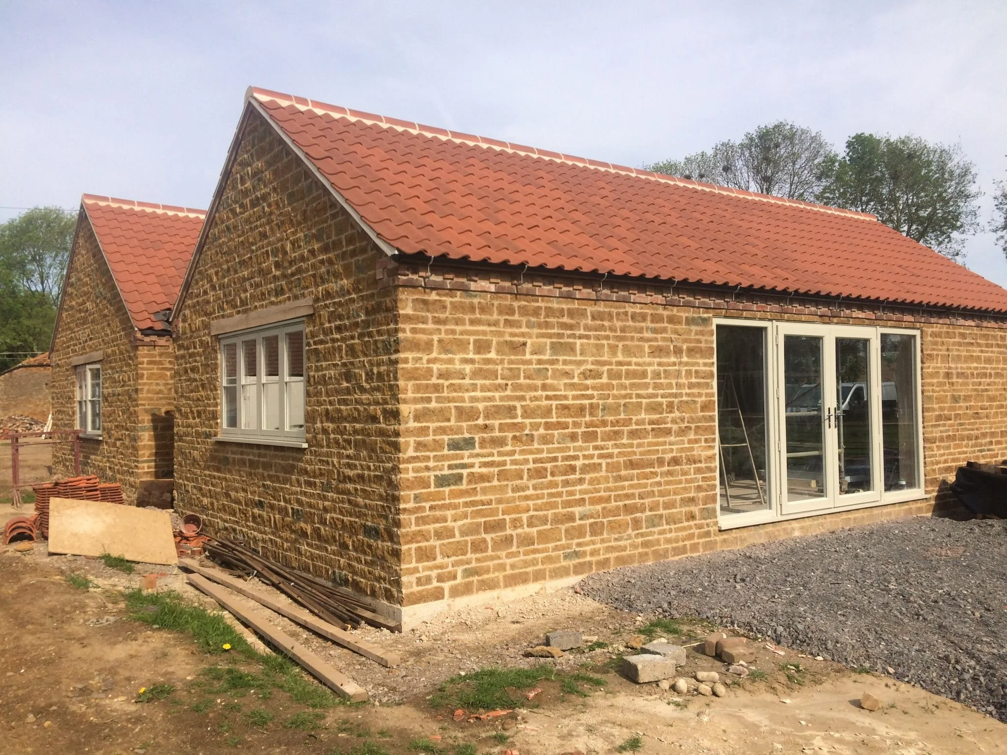 A brick house under construction with a red tiled roof and white-framed windows and doors, surrounded by construction materials and gravel.