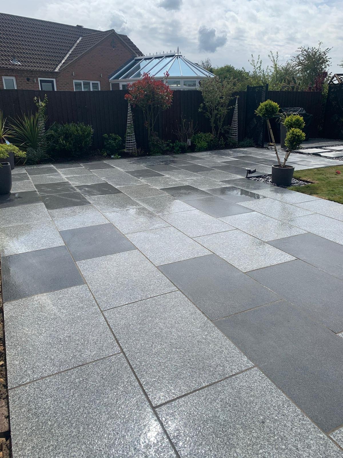 A paved backyard patio with gray and black tiles, surrounded by a black fence with plants and a small tree in a black pot, and a garden area with additional plants completed by local Grantham builder G East Builder.