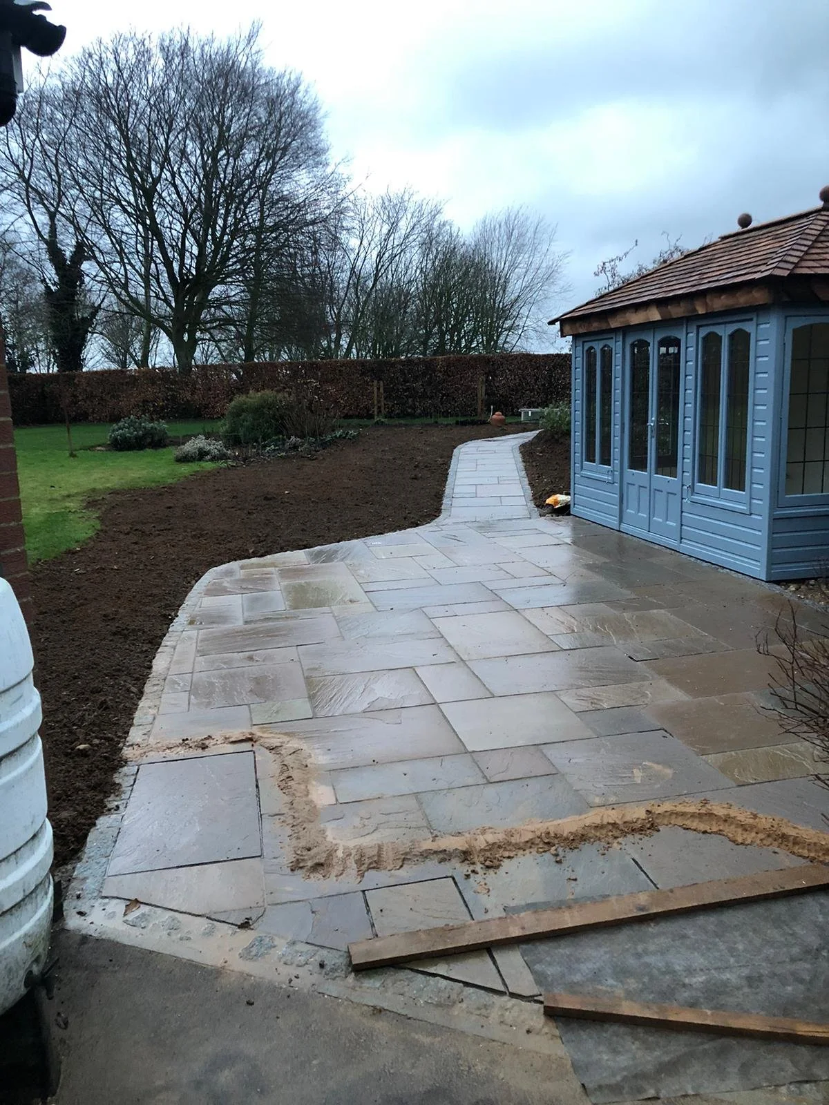 A newly paved stone patio with a winding pathway, adjacent to a blue garden shed, in a backyard with trees and a hedge, on a cloudy day.