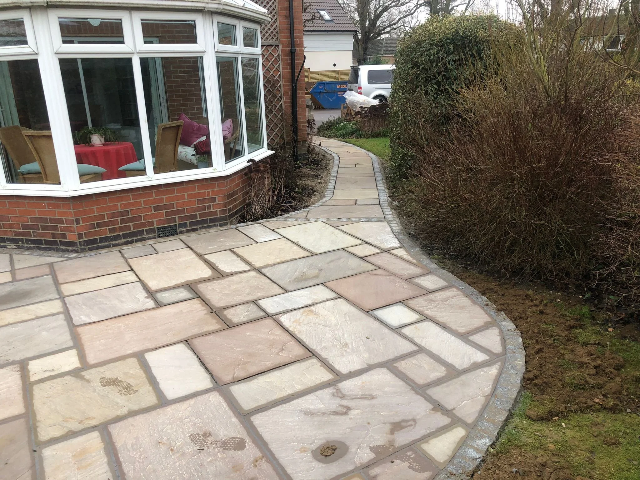 Curved stone paving walkway leading to the side yard of a house, with a bush border on the right and a brick house with large bay windows on the left.