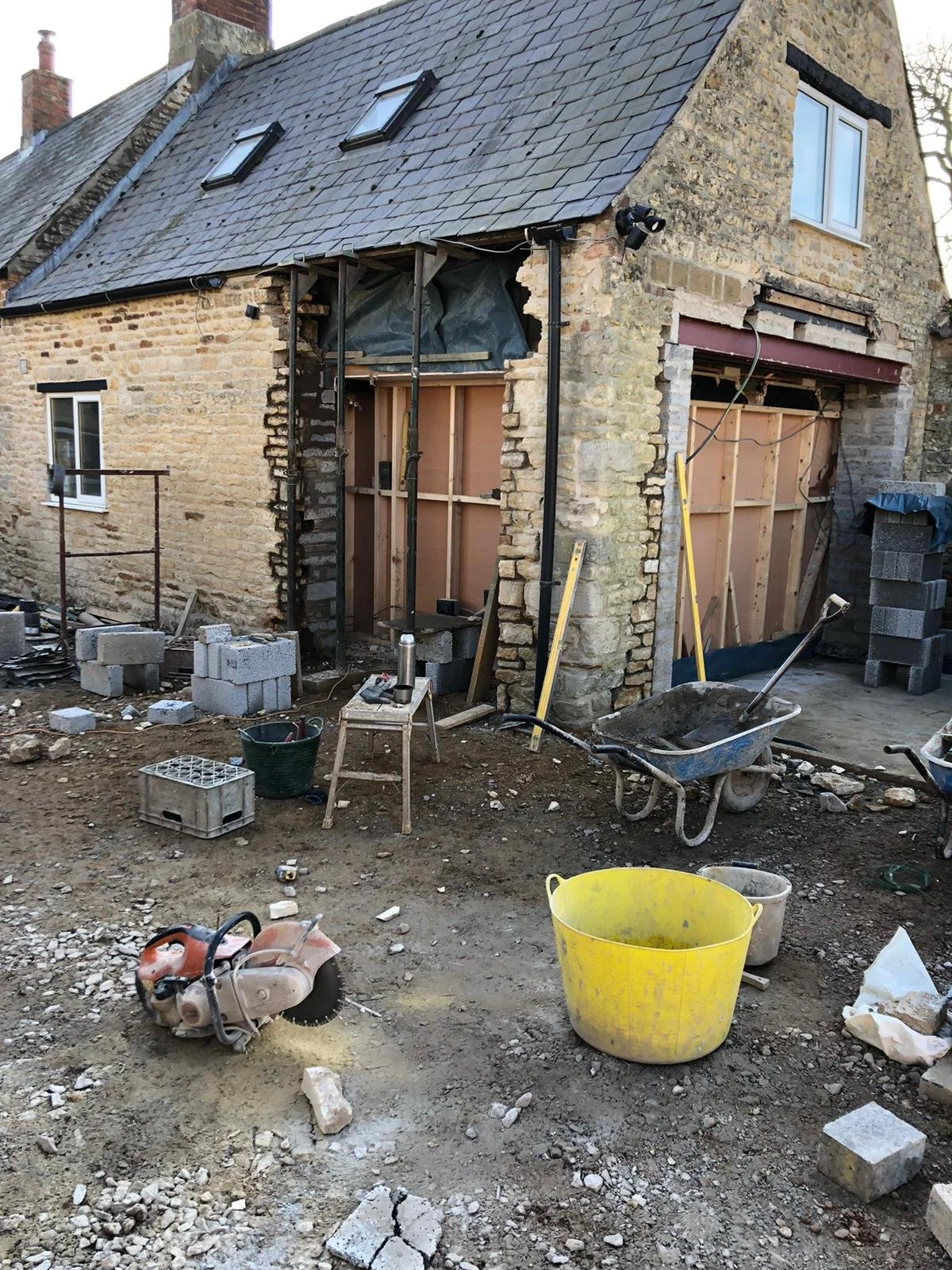 Construction site of a house renovation with building materials, tools, and equipment scattered around, including a saw, wheelbarrows, and large buckets, with part of the house's exterior walls and roof visible.