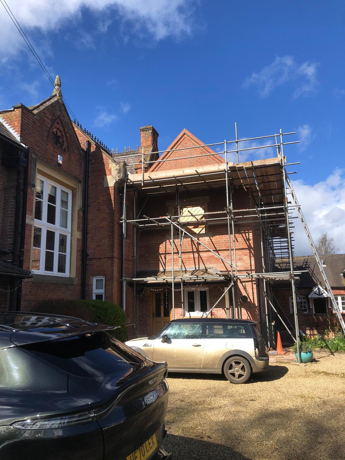A brick house under renovation with scaffolding set up around it, two cars parked in front, and a clear sky with some clouds.