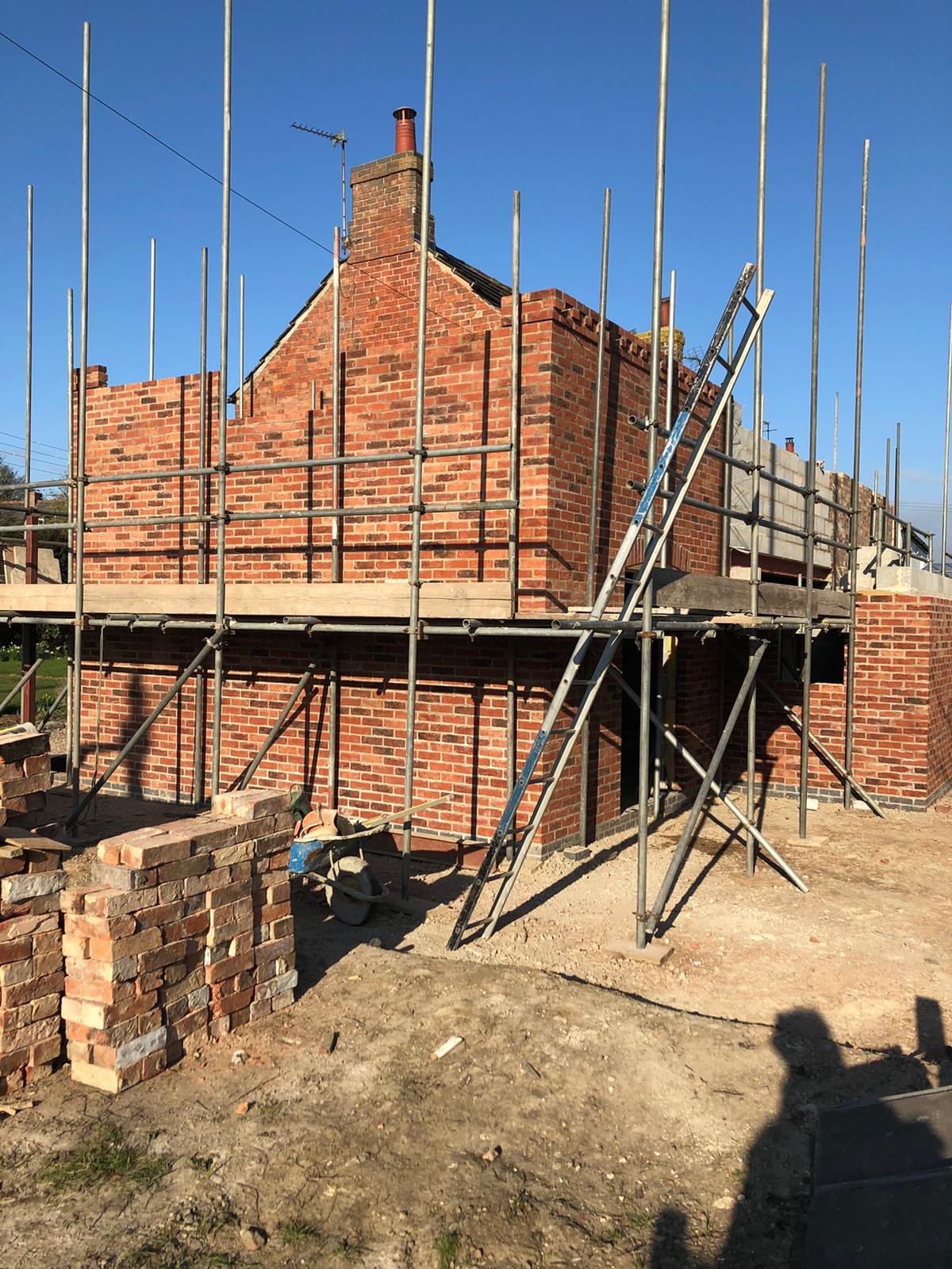 Construction site with scaffolding around a brick house under renovation or building in progress, with bricks and construction tools on ground, under clear blue sky.