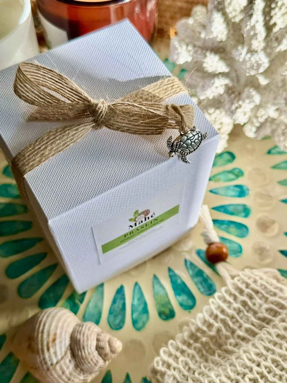 A white gift box wrapped with a beige string bow, decorated with a turtle charm, sitting on a yellow surface with green and blue leaf patterns. Surrounding items include seashells, a white knitted fabric, and a Christmas tree in the background.