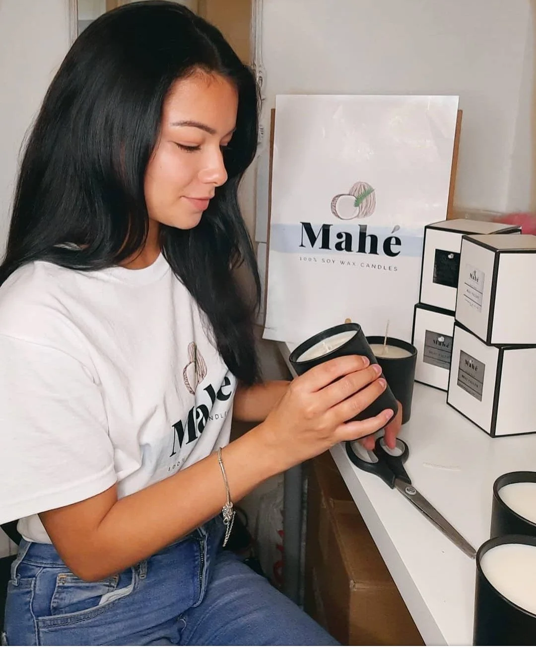 A woman with long black hair is holding a black candle container, carefully pouring or inspecting a candle wick. She is sitting at a white table with several black and white candles, a box cutter, and branded boxes of candles. In the background, there is a pack of candles and a sign that reads 'Mahé 100% Soy Wax Candles'.