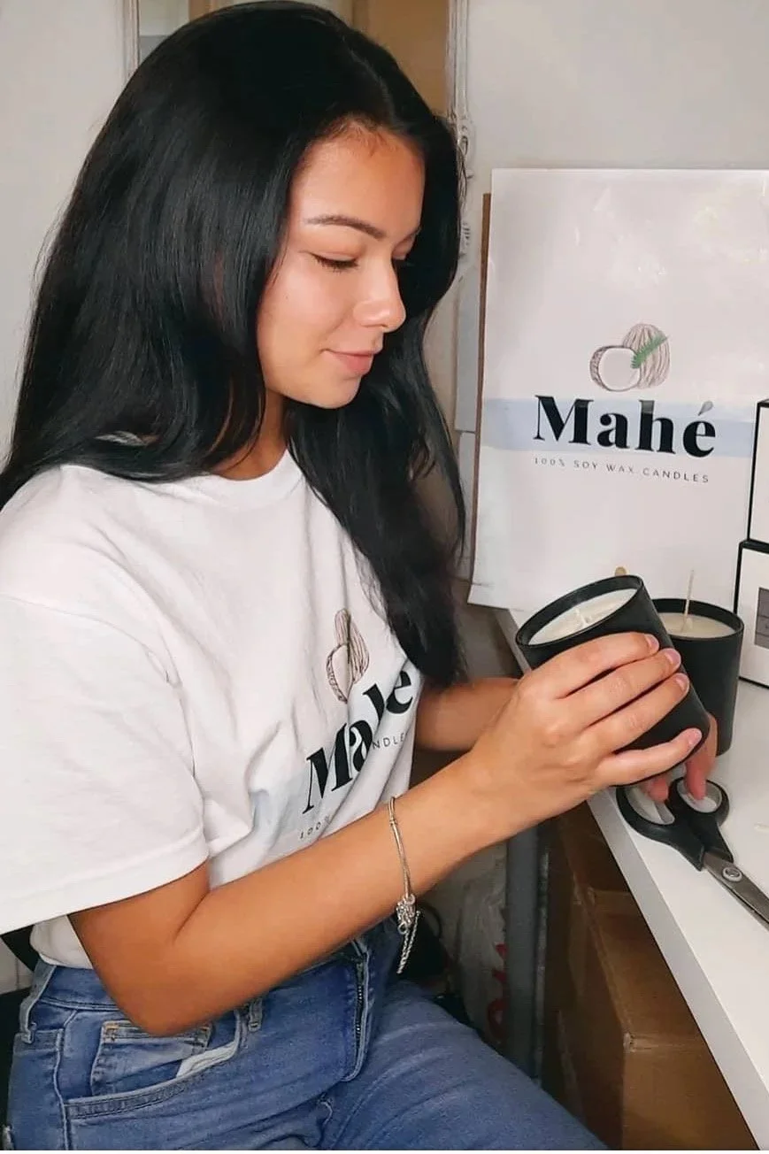 A woman with long black hair is holding a black candle container, sitting at a table with candles and a sign that reads 'Mahe 100% soy wax candles' behind her.