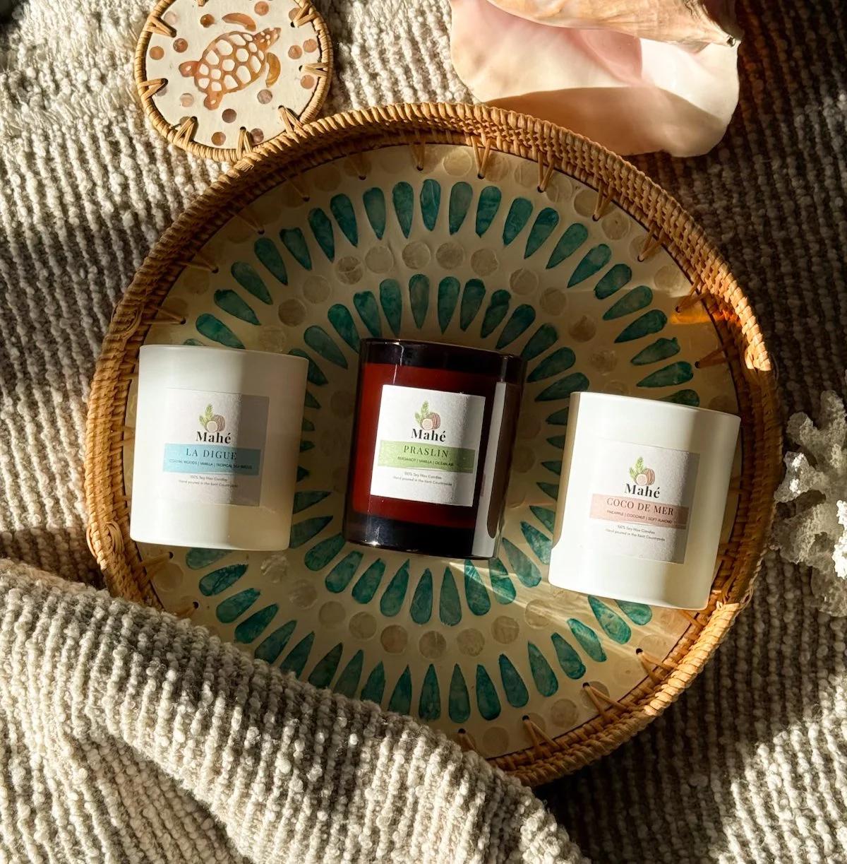 A decorative round woven tray holds three candles with labels; two are white and one is dark brown. The tray is on a textured beige fabric with a small decorative piece showing a turtle design at the top.