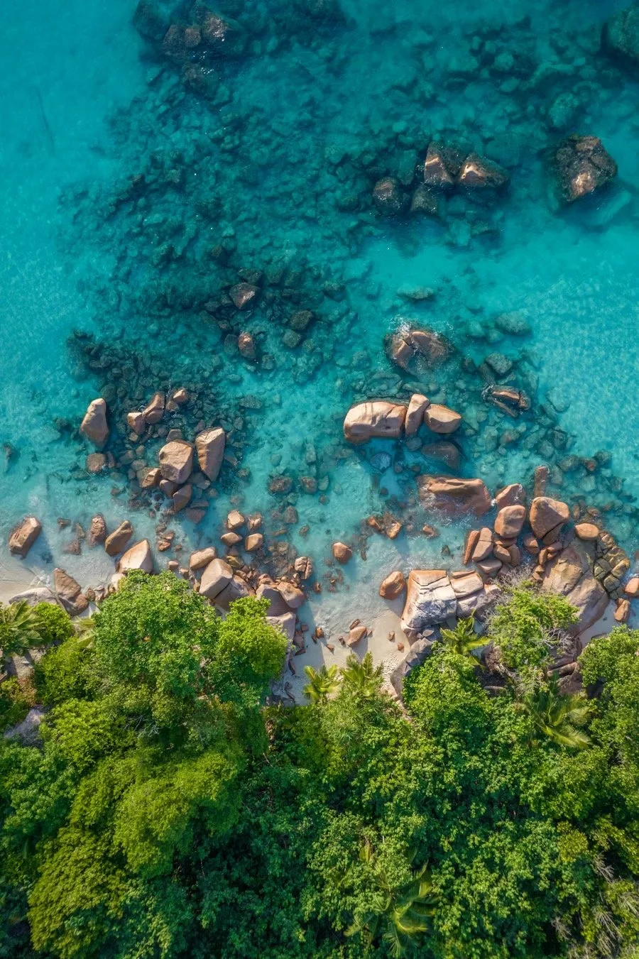 Aerial view of a tropical beach with crystal-clear turquoise water, large rocks, and green trees along the shoreline.
