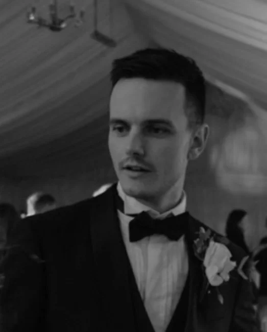 A young man in formal attire with a bow tie and a boutonniere, attending a social event or wedding reception.