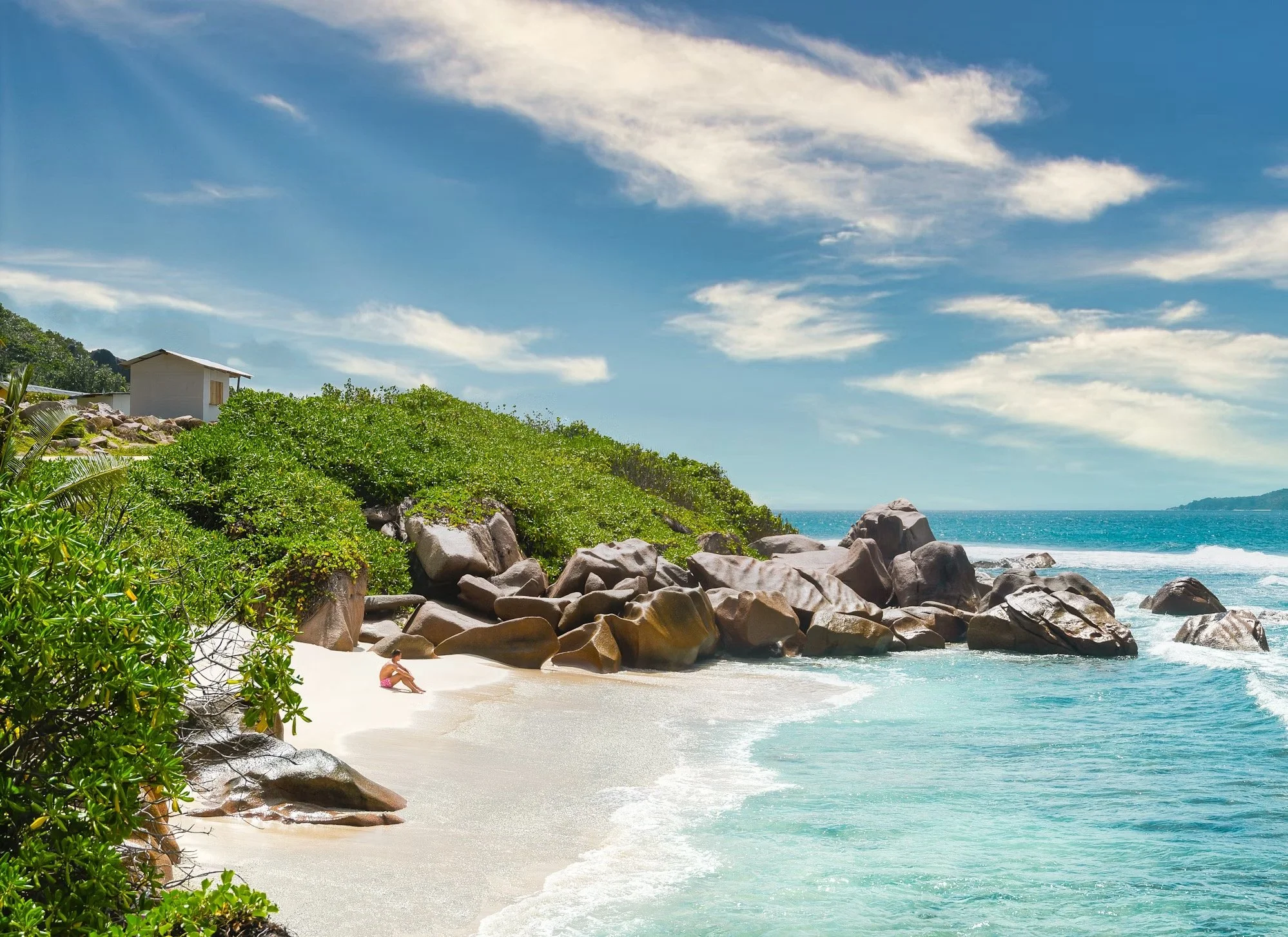 Tropical beach with white sand, green bushes, large rocks, and blue ocean under a partly cloudy sky.