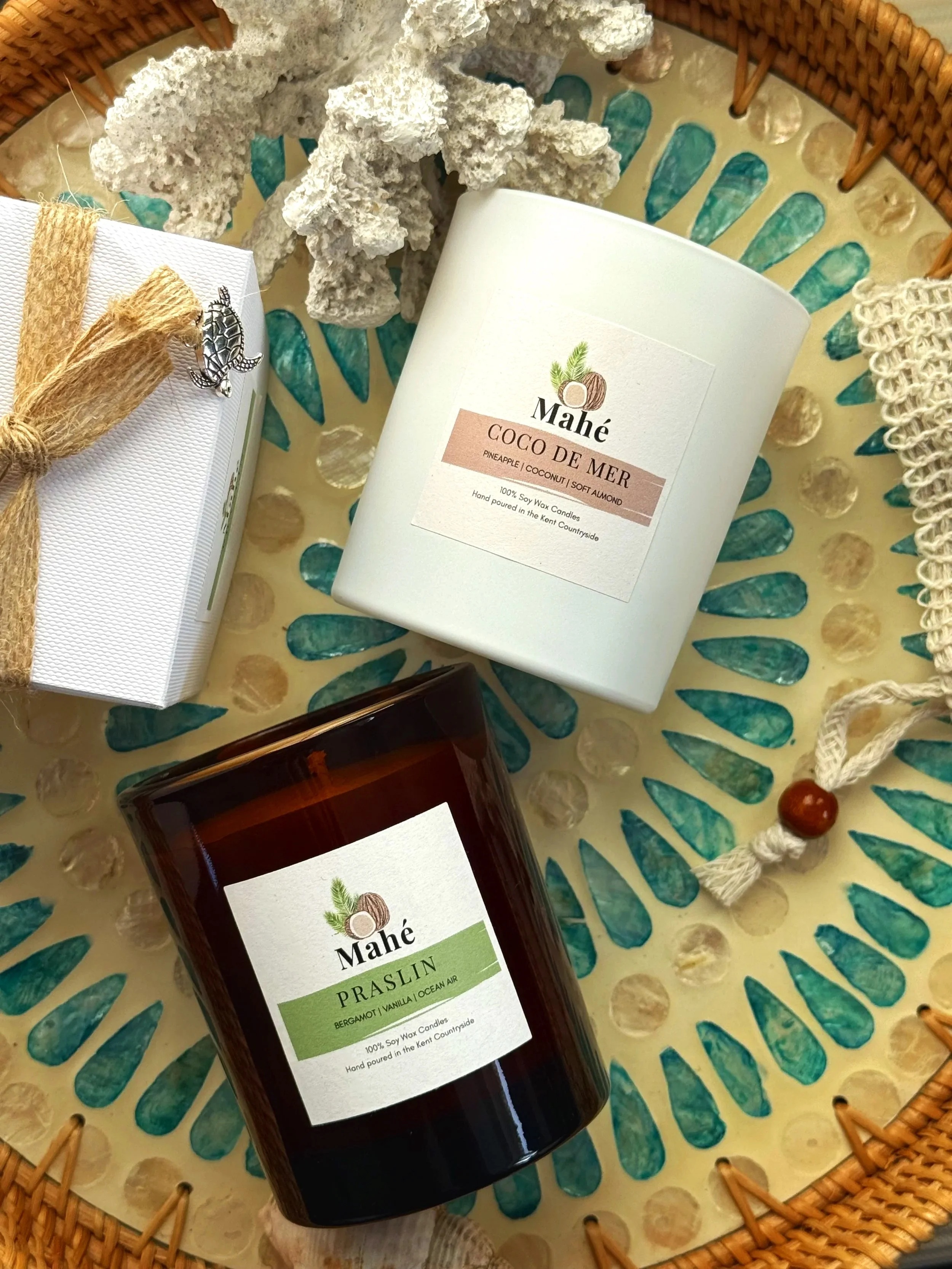 A basket containing two Mahé scented candles, one labeled 'Coco de Mer' and the other 'Prasin,' along with decorative coral and shells.