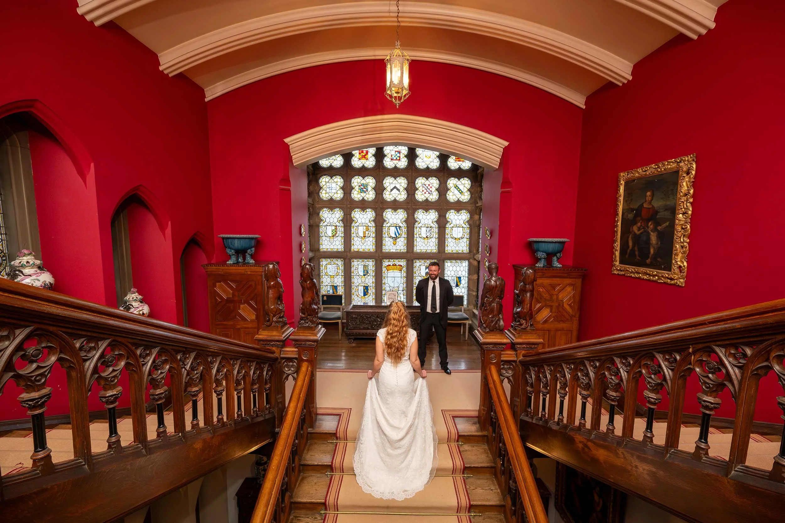 A bride and groom stand on the staircase of an elegant hall with red walls, wooden railings, stained glass windows, and ornate decorations.