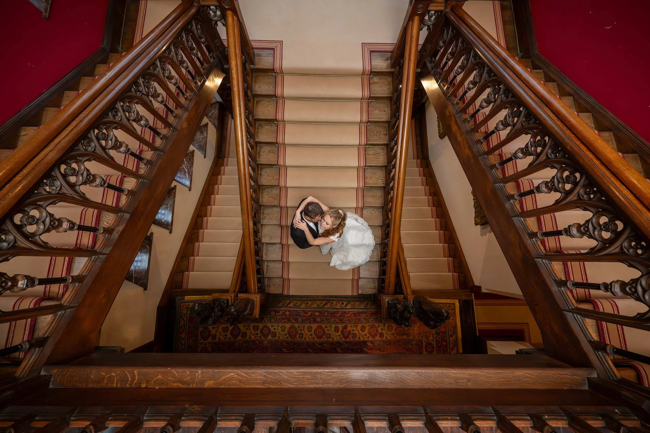 A bride and groom in wedding attire sharing a kiss at the top of a wooden staircase, viewed from above.