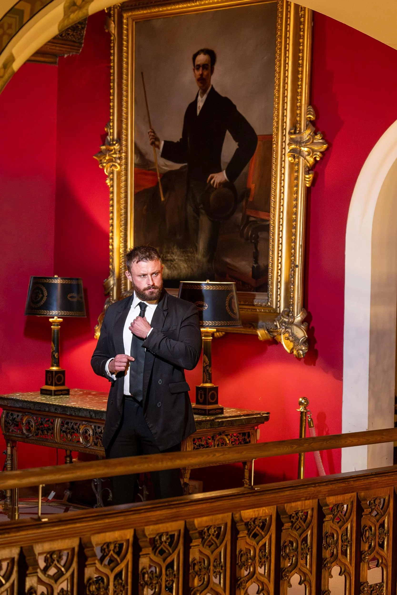 A man in a black suit and tie stands in front of a large, ornate portrait of a man holding a stick, with a red wall and decorative lamps and furniture in the background.