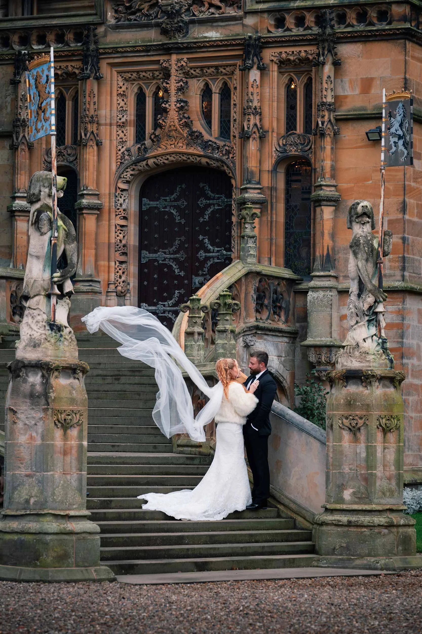 Bride and groom standing on stone steps in front of a gothic castle entrance, with ornate carvings and statues, celebrating wedding.