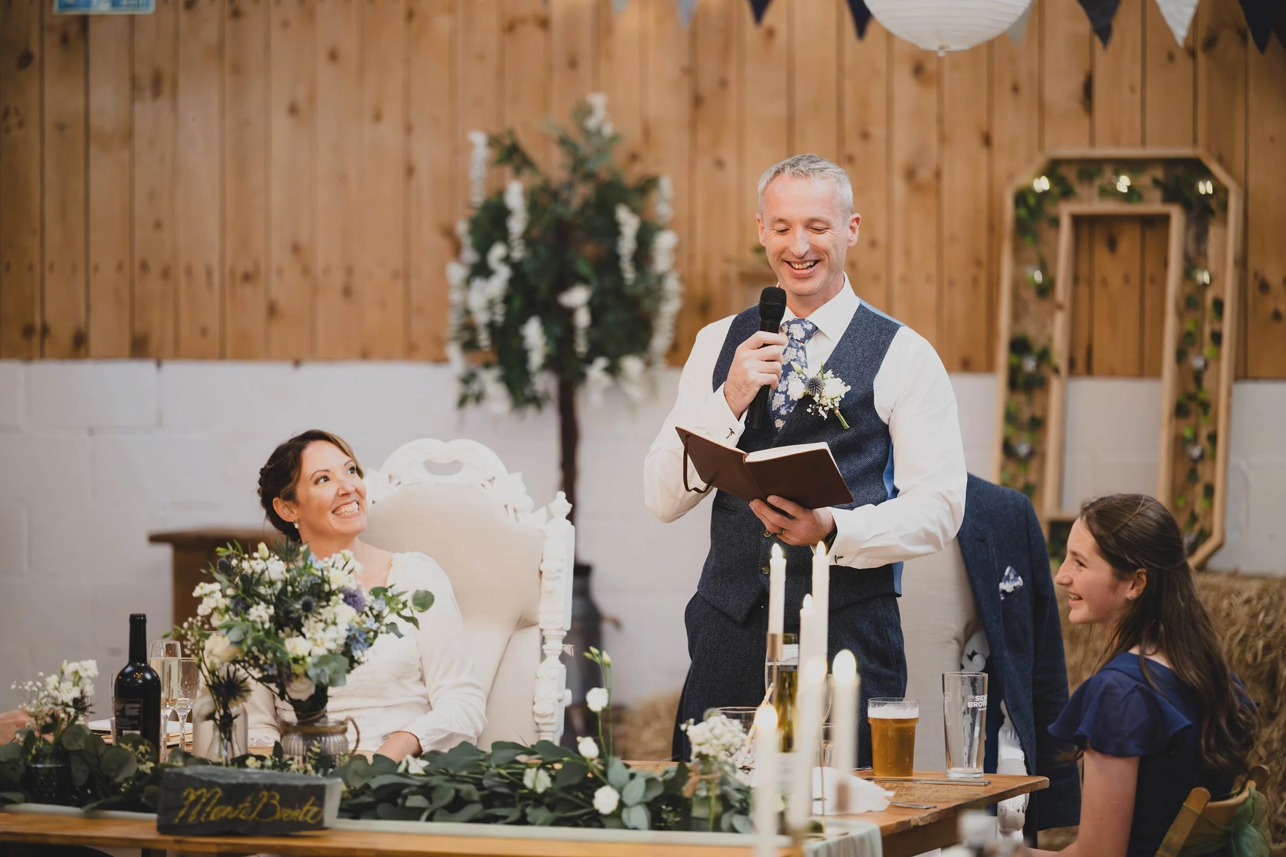 A man in a suit and vest giving a speech at a wedding reception, holding a microphone and reading from a book, with two women and a table decorated with flowers and candles in the background.