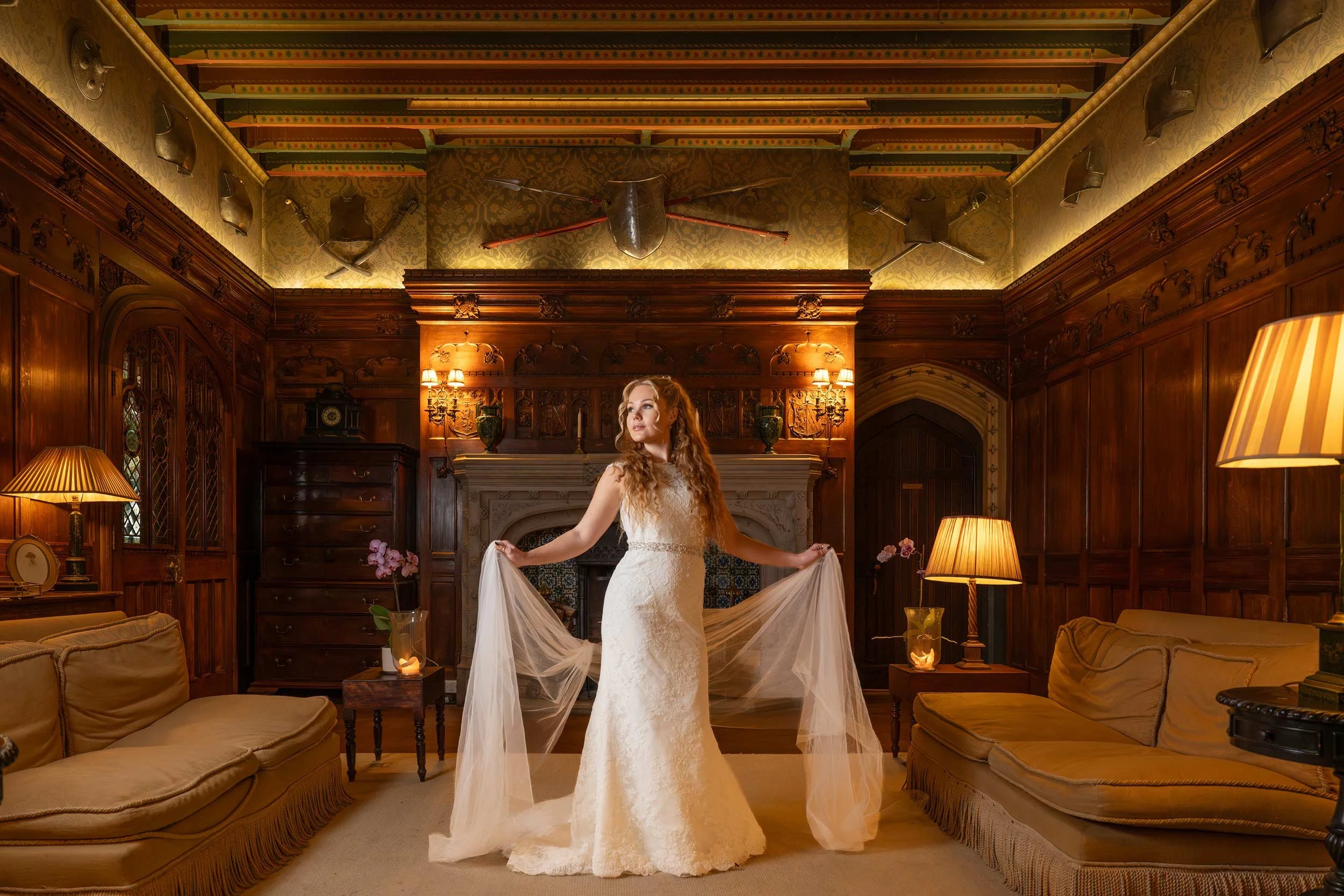 A woman in a white wedding dress standing in a richly decorated, wood-paneled room with a fireplace and lamps.