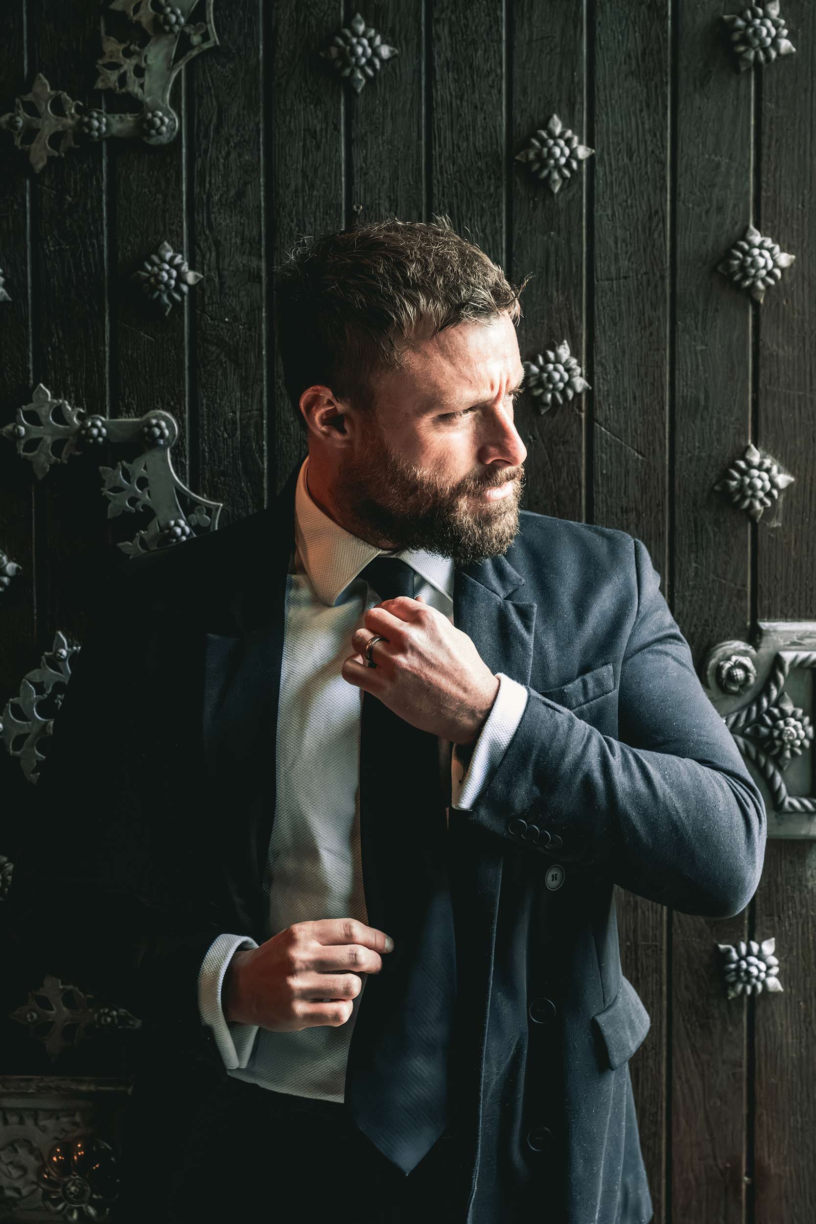 A man adjusting his tie while dressed in a dark suit in front of a black textured wall decorated with metallic floral and vine patterns.