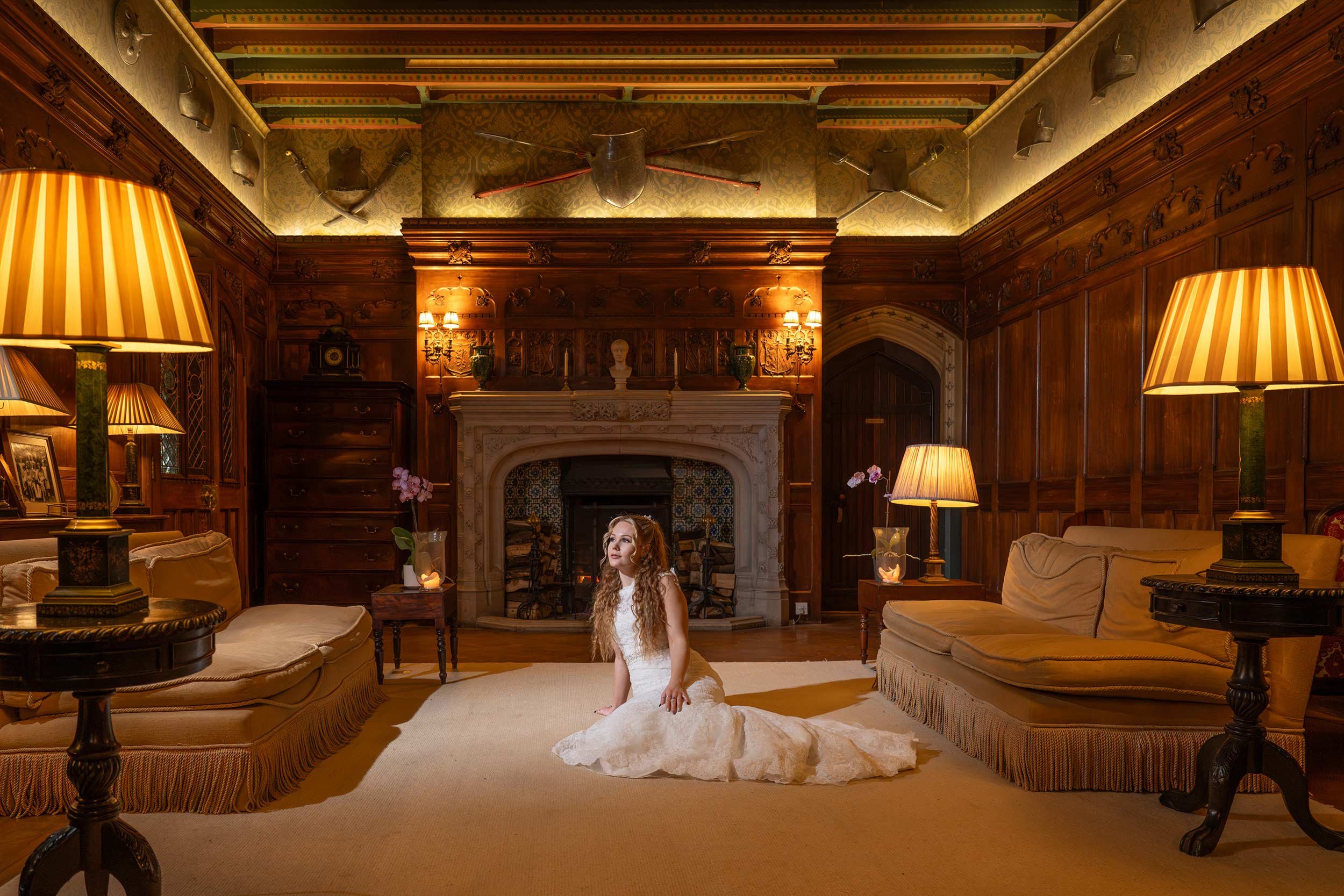 A woman in a white dress sitting on the floor in a richly decorated, wood-paneled living room with a fireplace. The room features ornate wooden furniture, lamps, and fireplaces with historic weaponry and decorative items on the walls and mantel.
