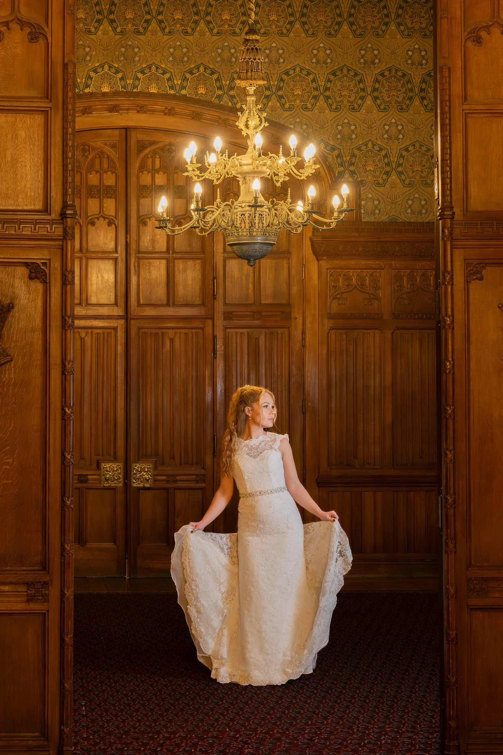 A woman in a white lace wedding dress standing in a wood-paneled room with a chandelier.
