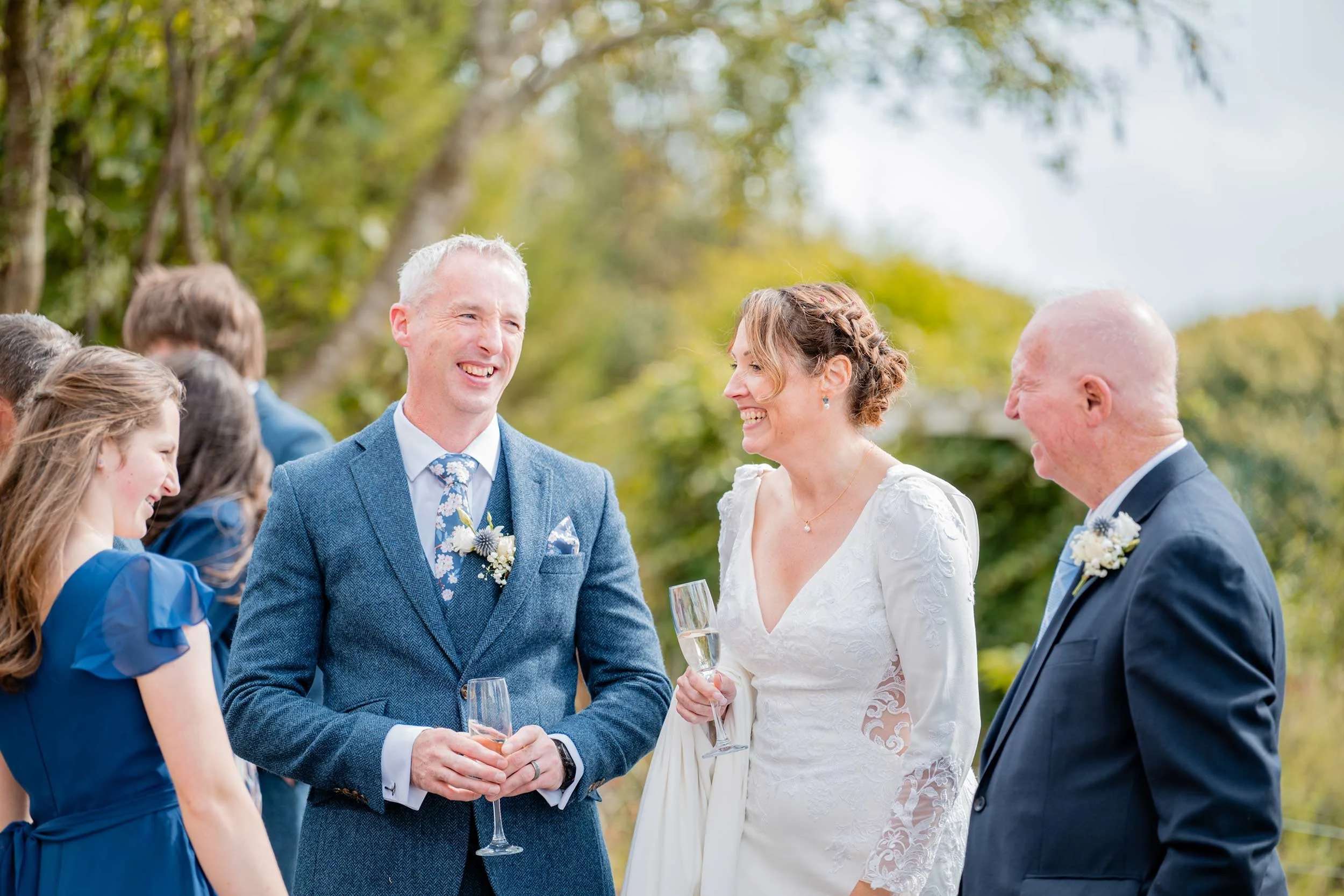 A wedding celebration outdoors with smiling bride, groom, and guests, holding champagne glasses, surrounded by green trees.