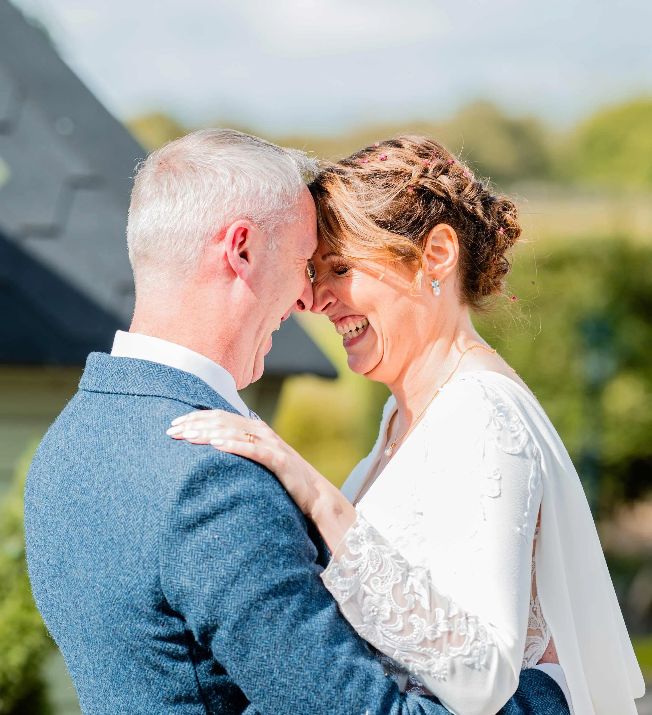A smiling couple, a man and a woman, are holding each other close with foreheads touching, outdoors on a sunny day. The woman is wearing a white dress with lace details, and the man is dressed in a blue suit.